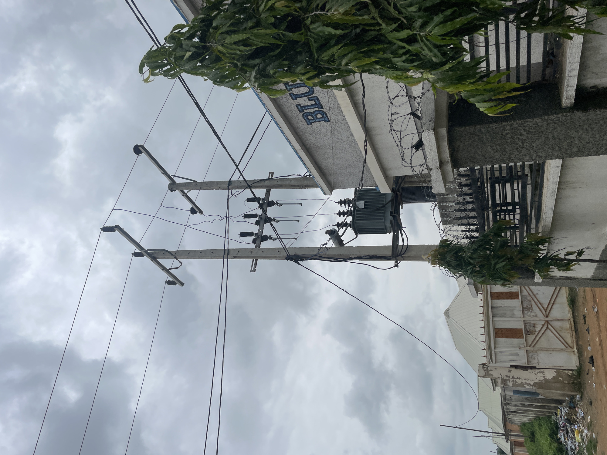 The image shows a street scene with overhead electrical wires, a transformer, and a building with the word "BLUE" visible on its facade, located in an urban environment in Unknown, Nigeria. A concrete utility pole with insulators and wires is prominent in the foreground, supporting several electrical lines. A large electrical transformer is mounted on the utility pole. Adjacent to the pole is a wall topped with barbed wire and a metal fence. A building with a light-colored, textured facade has the word "BLUE" inscribed in large, dark blue letters. To the left, a row of residential buildings with white, gabled roofs and gates can be seen. A pile of trash is visible on the ground in front of these houses. A tall, leafy tree partially obscures the right side of the frame. The sky is overcast with gray clouds.