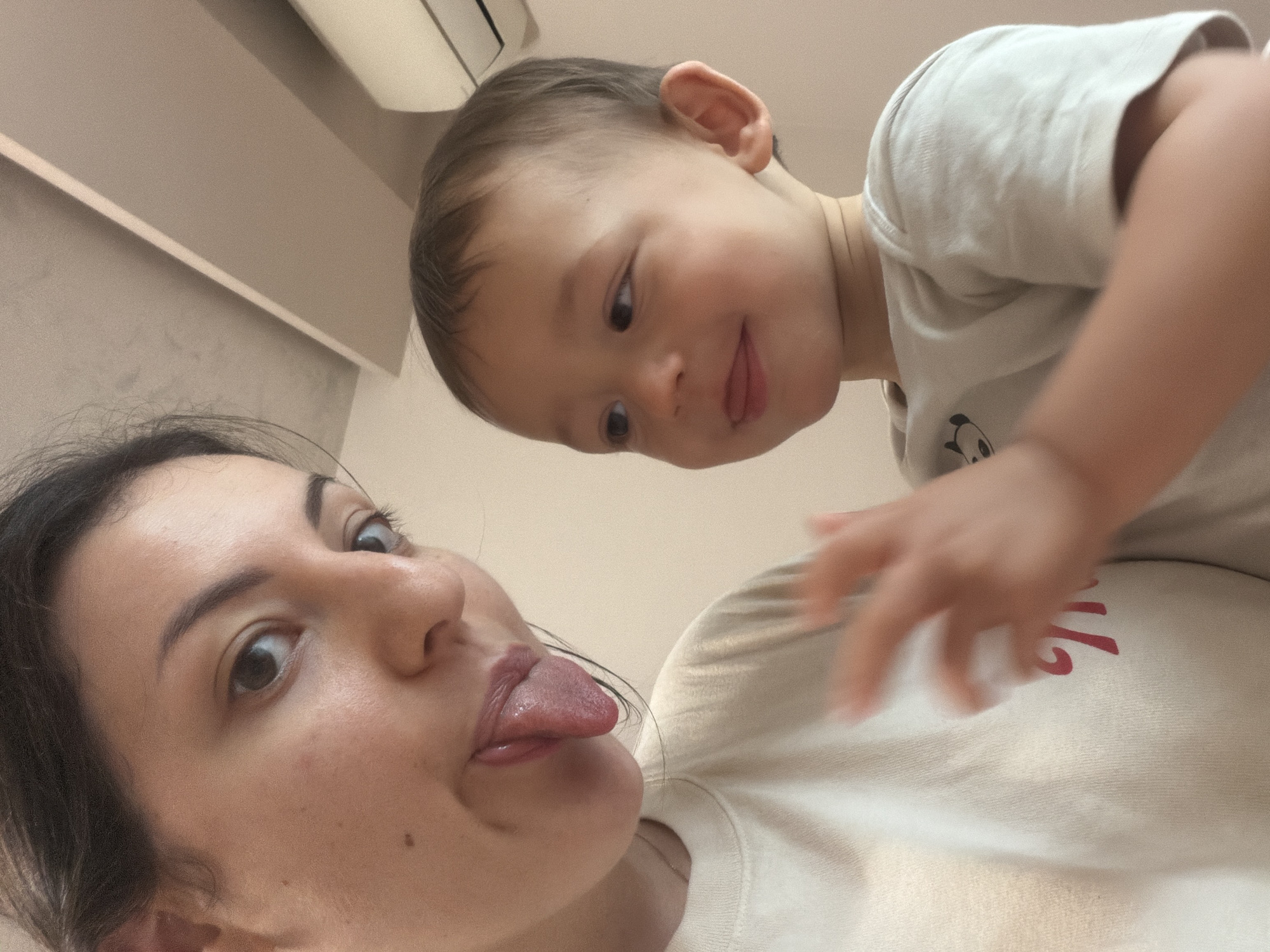 A low-angle selfie captures a woman and a young child, likely a toddler. The woman is in the foreground, and she's sticking her tongue out. The child, positioned above her, is also sticking out his tongue. The child's hand is slightly blurred and raised as if in motion towards the camera. They are indoors. The background is a wall and a ceiling fixture. This image does not appear to be from Plovdiv, Bulgaria.