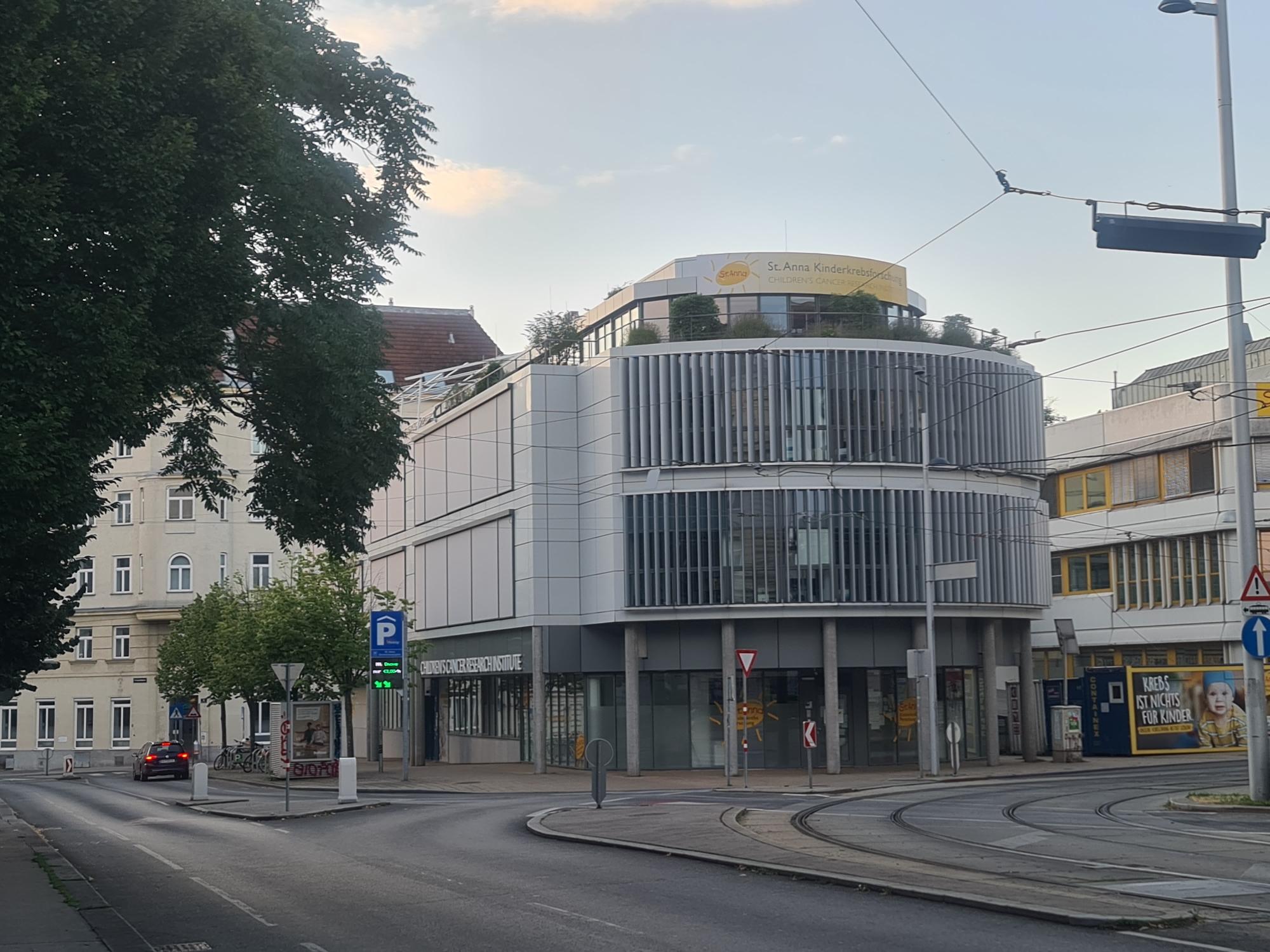 This is a medium-angle, eye-level, full shot of a street scene in Vienna, Austria. The photograph captures a modern, curved building with vertical louvers, identified by signage as the "St. Anna Kinderkrebsforschung" (St. Anna Children's Cancer Research). The building has large windows and a rounded upper section with greenery on its roof. In front of the building, there are tram tracks, traffic lights, and various street signs. A car is visible driving on the road, and several bicycles are parked near the entrance. The street is paved and appears to be an intersection with tram lines. To the left of the modern building is a more traditional, older building with multiple windows and an orange-tiled roof. The sky is partly cloudy, with the sun setting. A large billboard advertising "Krebs ist nichts für Kinder" (Cancer is not for children) is on the right side of the frame. The overall scene depicts a busy urban environment.