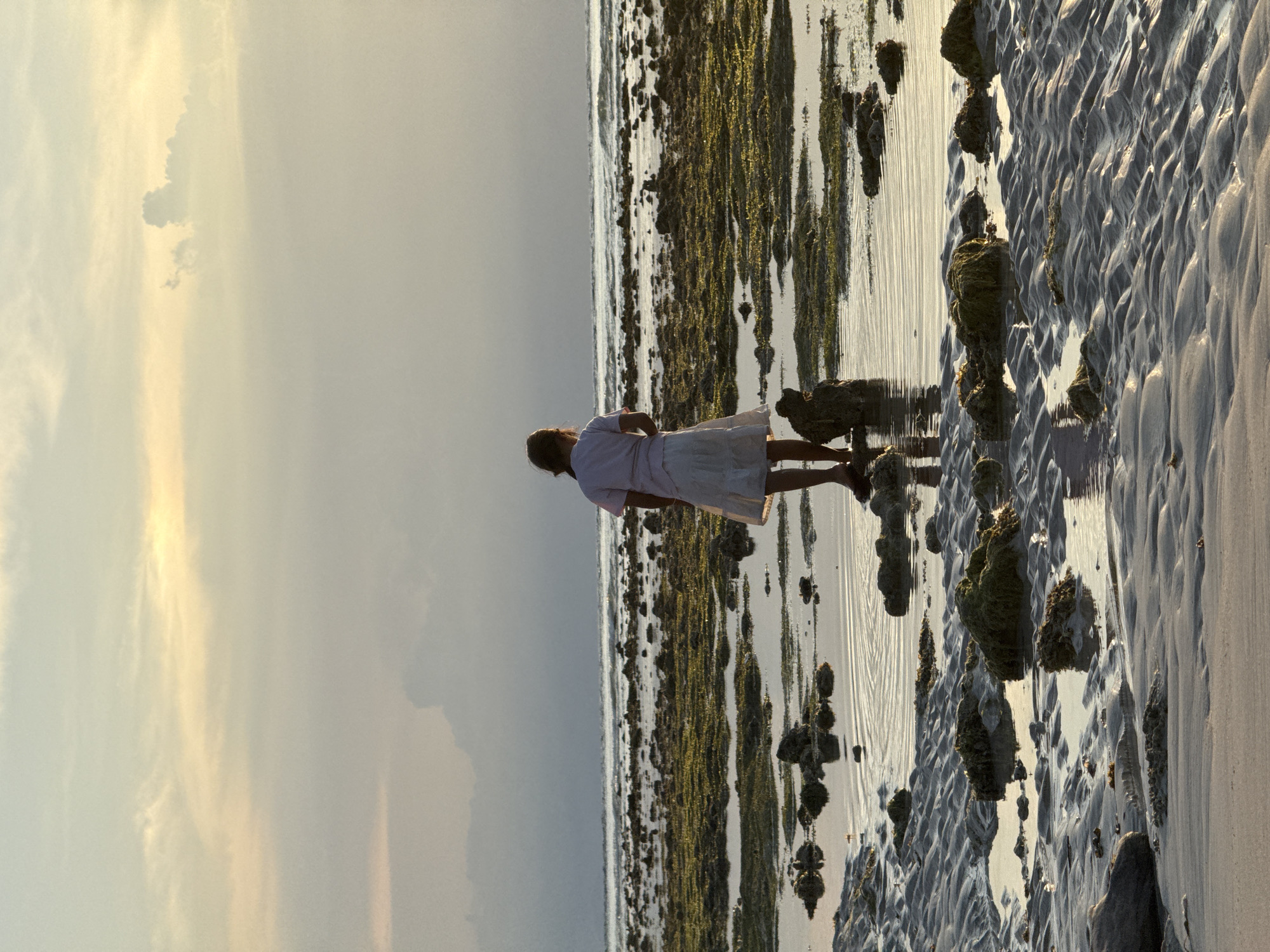 A woman in a light-colored dress stands on a beach at low tide, likely in the city of Unknown, Indonesia. She is facing away from the viewer and towards the receding water line. The beach is textured with wave patterns in the sand. Rocks and seaweed are scattered across the exposed seabed. The sky is overcast with patches of lighter clouds.