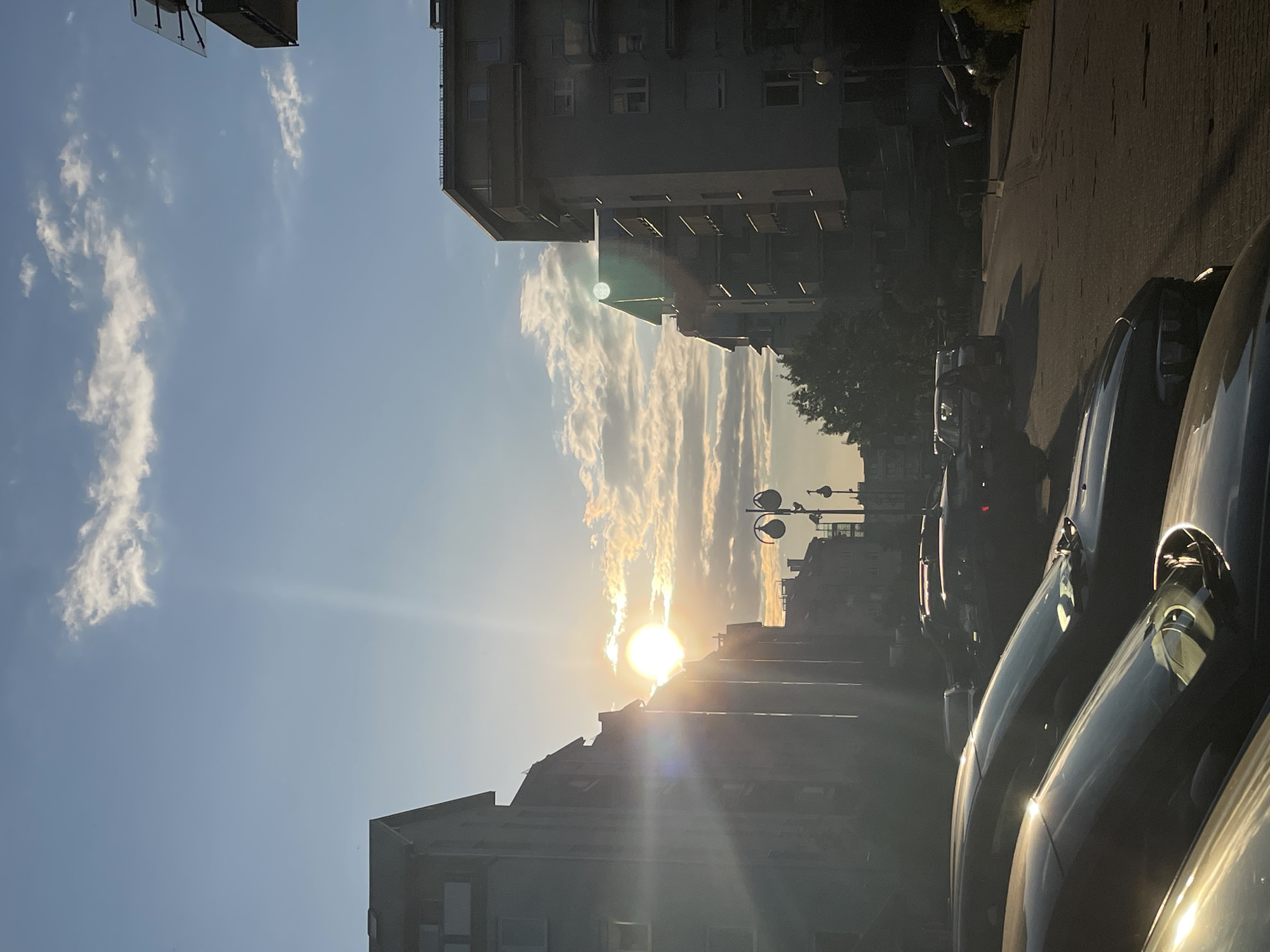 The image depicts a city street scene at sunrise or sunset. The sun is positioned low on the horizon, partially obscured by buildings, casting a bright glare and long shadows. There are buildings on both sides of the street, suggesting an urban or suburban setting. Parked cars are visible along the right side of the frame, filling a significant portion of the foreground. The sky is blue with streaks of clouds that appear to be illuminated by the sun. Streetlights are visible in the distance. The buildings are unremarkable, appearing to be residential or commercial structures. The overall impression is one of an ordinary urban environment at the beginning or end of the day.