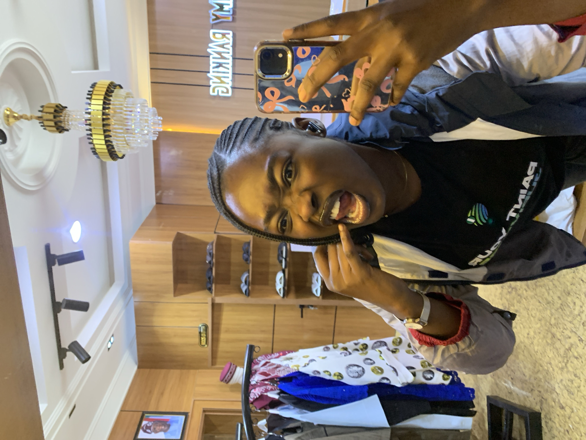 In a well-lit interior, likely a retail space in Gura Top, Nigeria, a young Black woman is taking a selfie in a mirror. She is wearing a black t-shirt with a green and white logo, and a grey and blue jacket with red trim. Her hair is styled in thin braids, and she has a playful expression, sticking out her tongue and gesturing with her hand towards her mouth, as if to emphasize her teeth. Her left wrist is adorned with a silver watch.
The background reveals a store environment. A wooden wall features shelving displaying pairs of footwear, possibly sandals. To the left, clothing is hung on a rack, including patterned shirts and a traditional cap. Above, a contemporary track lighting system is installed on the ceiling, and a multi-tiered crystal chandelier hangs prominently. In the reflection, a neon sign spelling "MY BANKING" is visible on a wooden panel. A framed portrait of a Black man in formal attire hangs on the far left wall. The flooring appears to be a textured material.