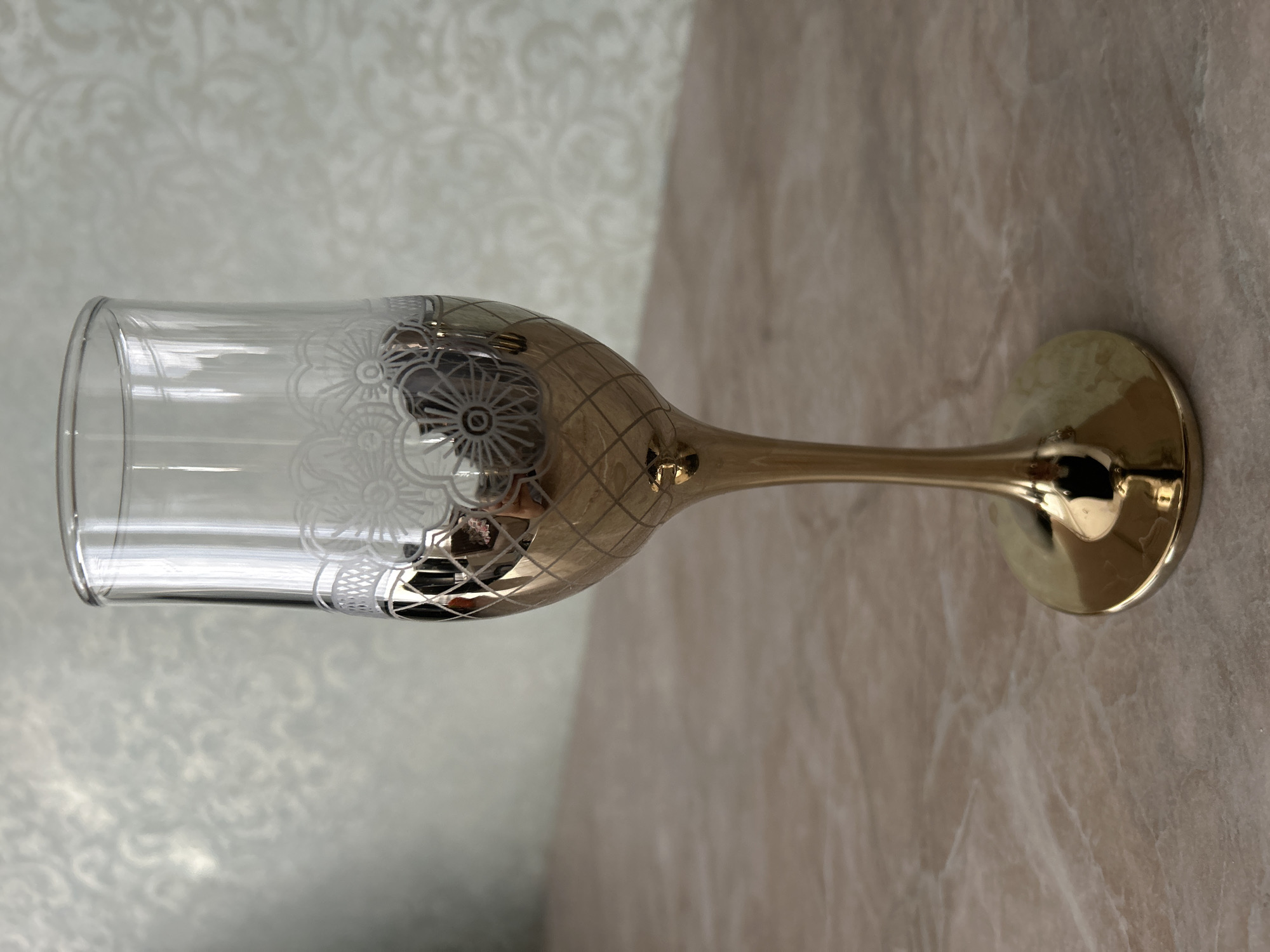 A close-up, eye-level shot shows a single, empty champagne flute with a gold-colored stem and base. The glass bowl is clear and features a decorative band of white floral etchings near its rim. The lower portion of the bowl is also gold-colored and has a diamond-shaped pattern. The flute stands on a beige, marble-like surface, with a textured, light-colored wall in the background. The overall lighting is soft and natural, creating subtle reflections on the metallic surfaces of the glass. The location is not discernible from the image.