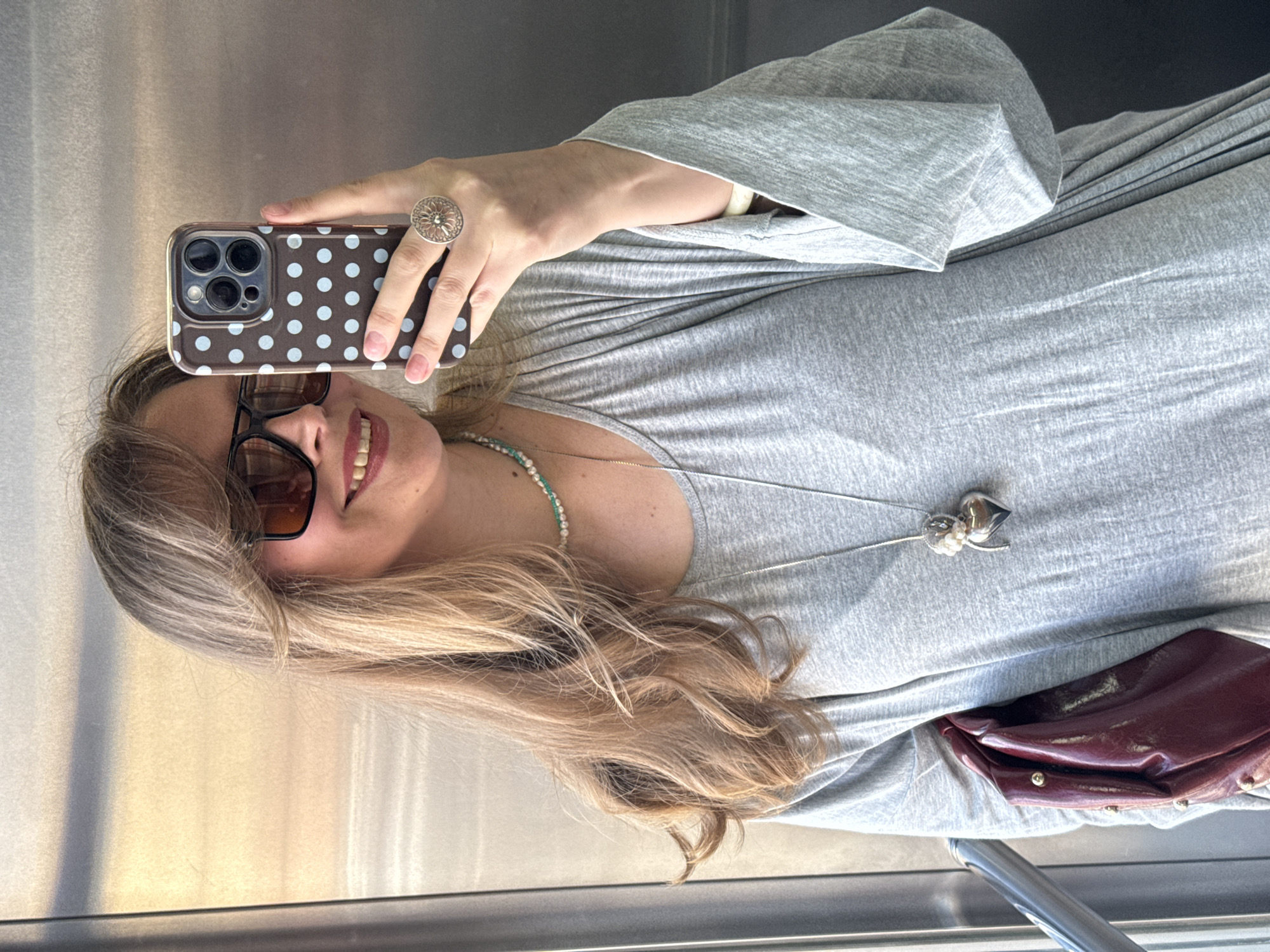 A woman takes a mirror selfie in an elevator. She has long blonde hair with bangs and is wearing sunglasses, a grey dress, a pearl necklace, and a silver necklace with a heart pendant. She is also wearing a large silver ring. She is holding a phone with a polka dot case. A maroon bag is visible on her left. The elevator walls are metallic. The location is not specified as being in Izhevsk, Russia.