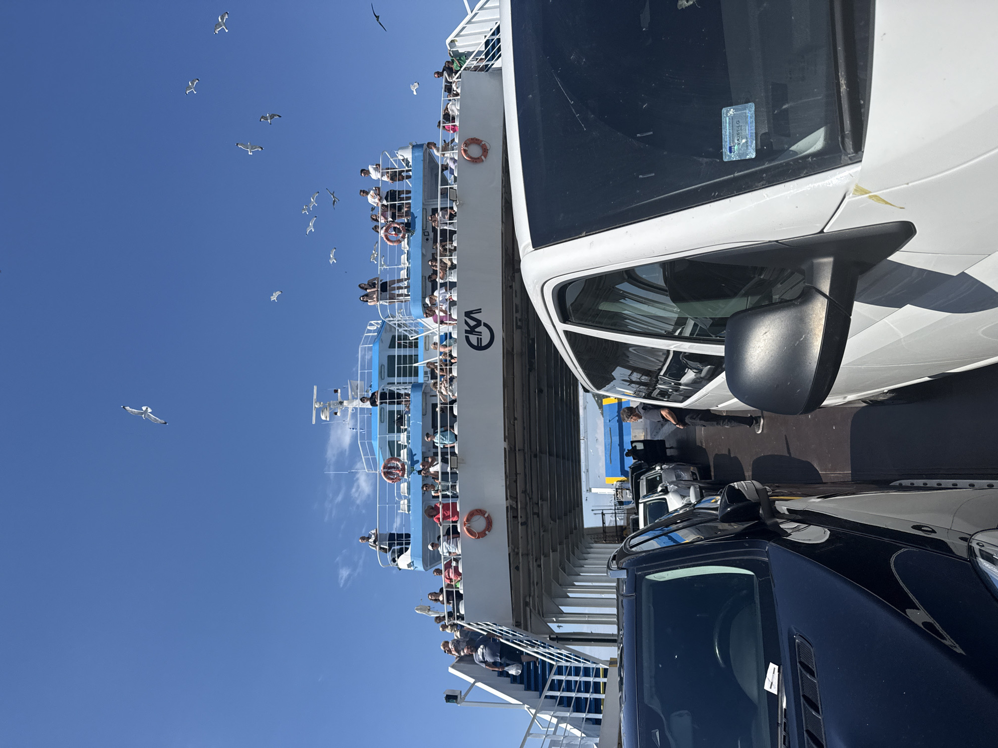 This is a medium shot of a ferry in Kavala, Greece, carrying cars and passengers. The ferry has multiple decks with people standing and observing the surroundings. Several cars are lined up on the ferry deck. In the foreground, a black car is visible on the left, and a white van is on the right. The sky is clear blue with several seagulls flying around the ferry. The ferry has the logo "EKA" visible on its side. The overall scene depicts a sunny day with a ferry journey underway.