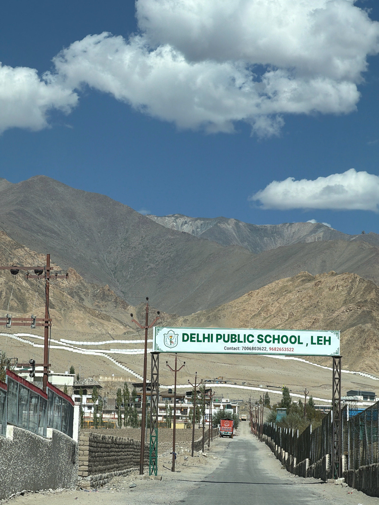 The image captures a street scene in Leh, India, under a partly cloudy blue sky. In the background, rugged, arid mountains dominate the landscape, their slopes marked by lighter-colored terraced structures that ascend the hillsides. A prominent green and white sign arching over the road announces "DELHI PUBLIC SCHOOL, LEH" with contact numbers displayed below.
In the mid-ground, a paved road leads into the distance, bordered by various buildings with flat roofs, characteristic of the region's architecture, interspersed with tall, slender trees. Power lines and utility poles line the sides of the road. A red truck with "CHALLENGER" written on its side is visible on the road. On the left side of the image, a stone wall with a metal fence runs alongside the road, while on the right, a similar fence is present. The ground is a mix of dirt and loose stones. A few individuals are sparsely visible in the scene, including one person near the right edge of the image. The overall environment suggests a bright, sunny day in a mountainous region.