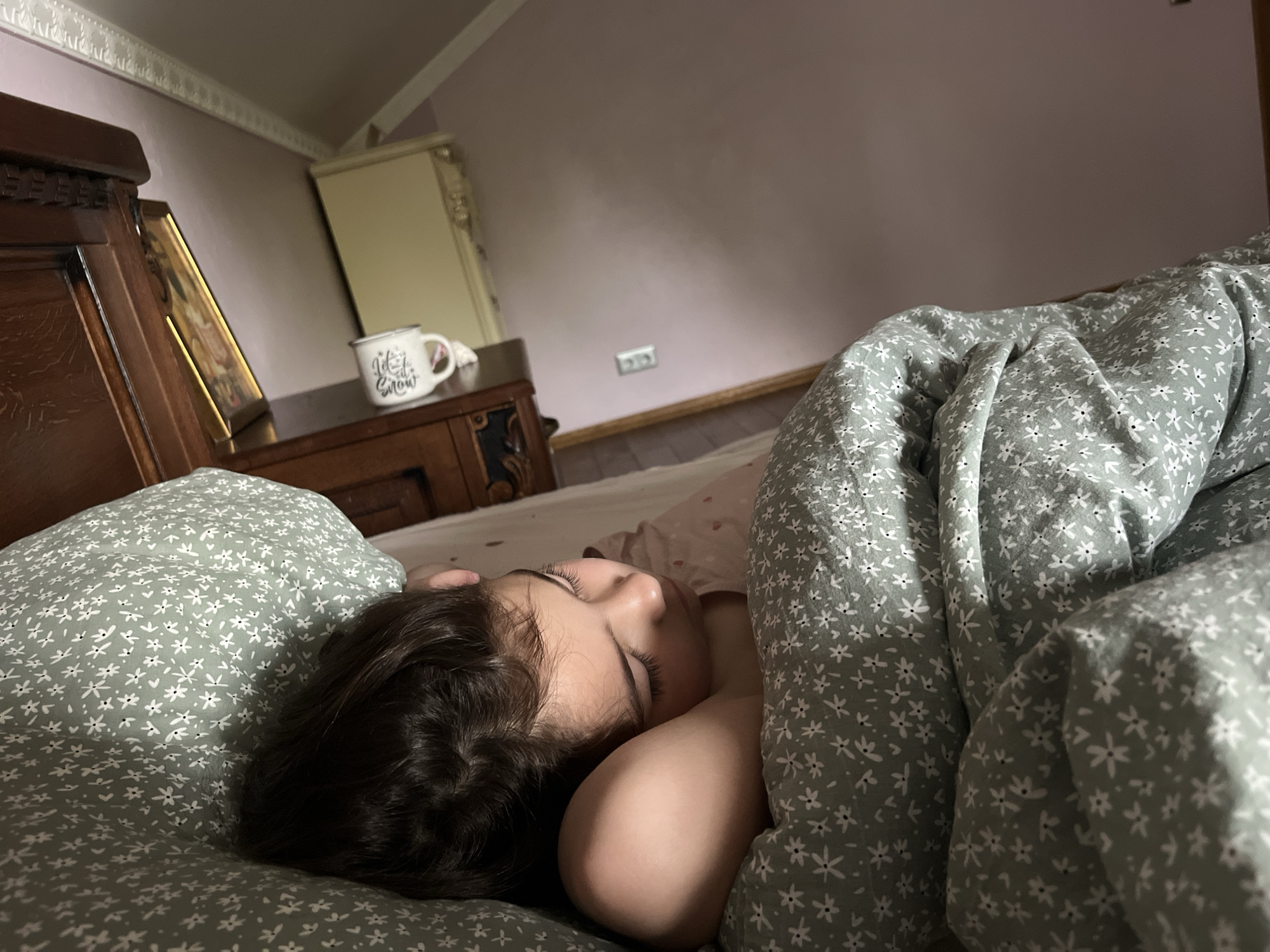 A young girl sleeps peacefully in a bed, her head resting on a pillow. She is covered by a floral duvet. The room appears to be a bedroom, with a wooden bedside table and a wardrobe visible in the background. The overall ambiance is one of quiet repose.
