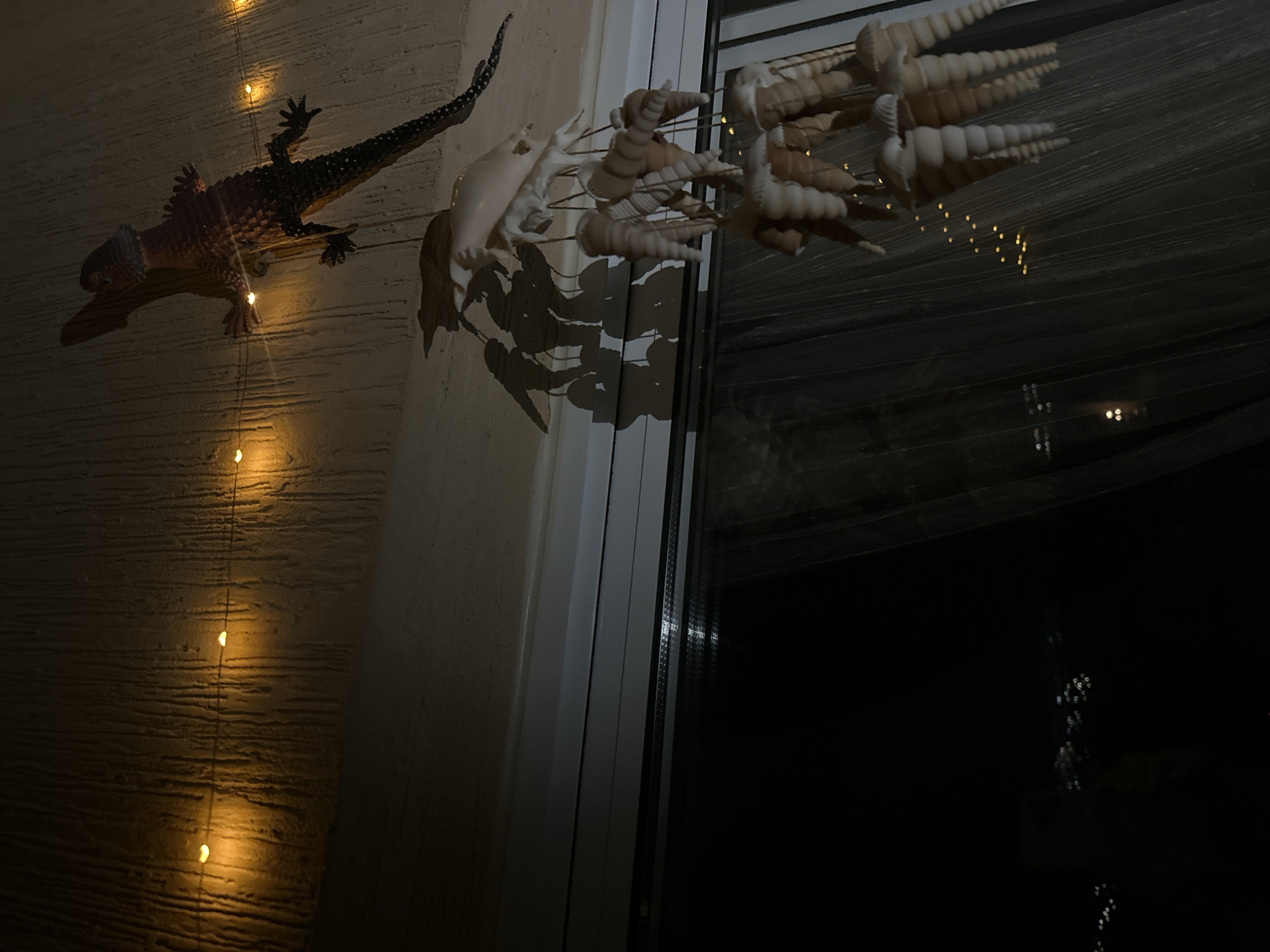 A decorative lizard figurine is affixed to a textured ceiling, illuminated by a string of warm, yellow fairy lights. Below the lizard, a shell wind chime hangs, its conical shells creating a cascading effect. The scene is set indoors, with a window visible in the lower half of the frame, revealing a dark, possibly nighttime exterior with faint, blurred lights. The overall ambiance is dim and atmospheric. The location is specified as Peyia, Cyprus.