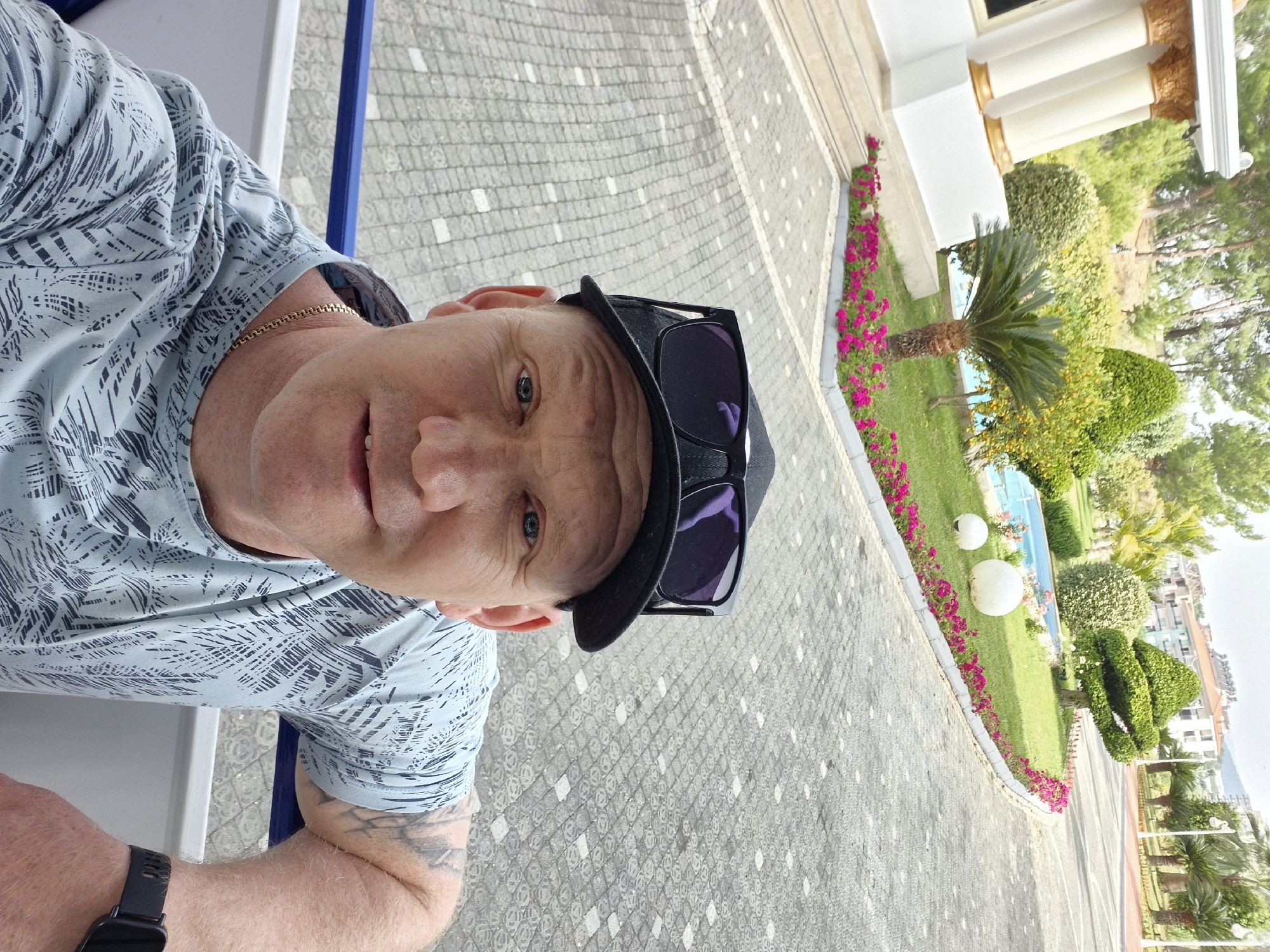 The image shows a man taking a selfie while on vacation. The man is wearing a light-colored patterned shirt, a gold chain, a black baseball cap with sunglasses perched on top, and a smart watch on his left wrist. He appears to be sitting on a white and blue chair. In the background, there's a paved walkway with a diamond pattern leading toward a building with white pillars. A meticulously maintained garden borders the walkway, featuring pink flowers, palm trees, and creatively shaped bushes. In the distance, there appears to be a swimming pool. Given the presence of palm trees, the architecture of the building, and the overall style of the landscape, it is likely that the location is in the city of Alanya, Turkey.