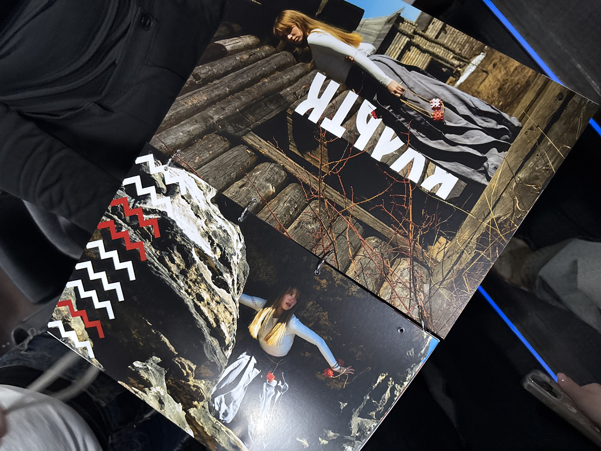 The image shows two overlapping pages of a book or magazine, likely a fashion spread, resting on a dark surface. The top page features a woman in a long grey dress and white top, lying against a wooden structure resembling a log cabin. The Cyrillic word "КУЛЬТ" is prominently displayed in white letters. The environment around the woman appears to be a rustic, possibly rural setting with bare branches.
The page underneath features another woman in a similar outfit, posed amidst a rocky outcrop. The foreground has a pattern of white and red zig-zags along the edge. The environment is a rock face.
The photo was taken in Perm, Russia.
