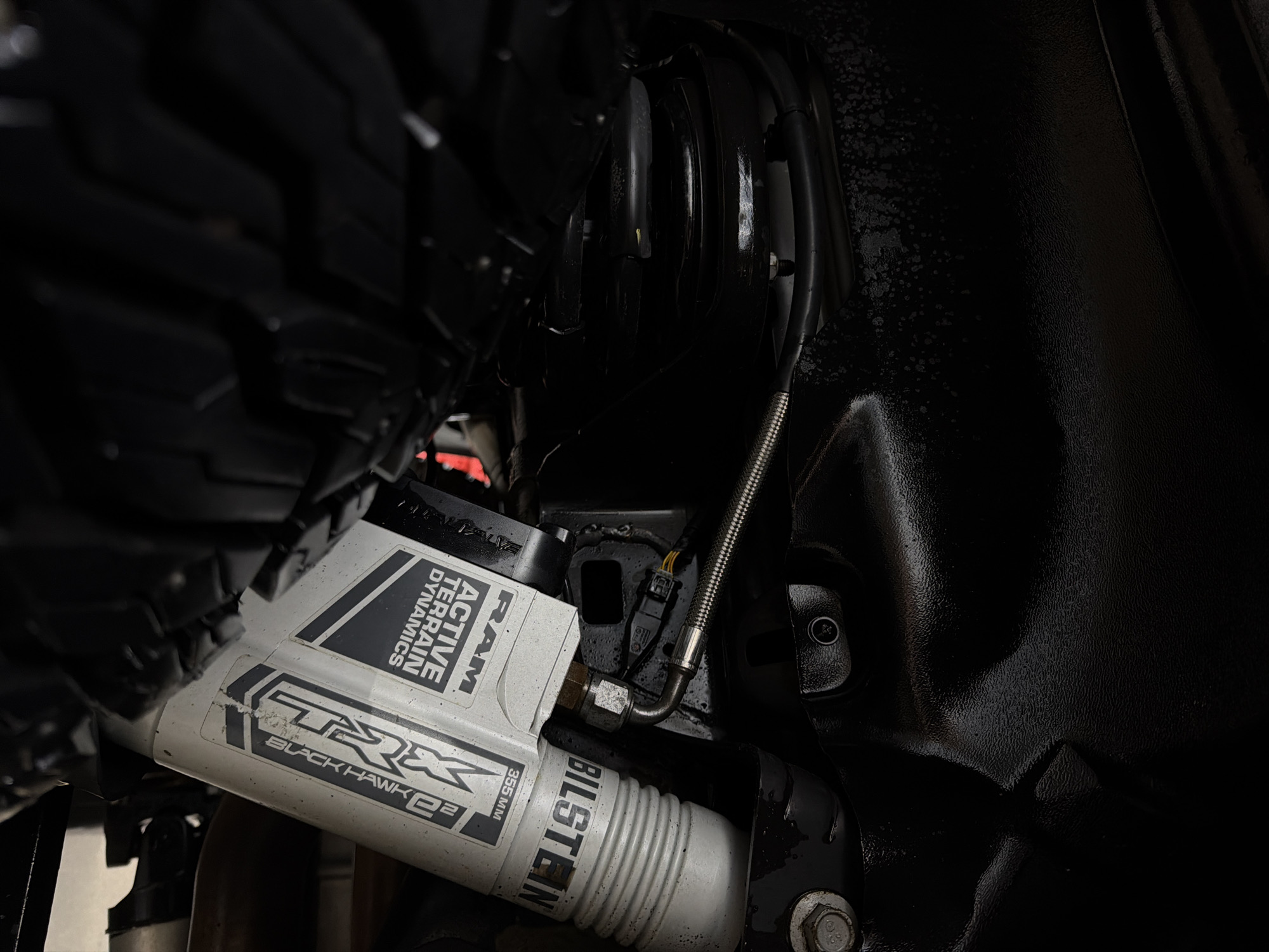 The image shows a close-up of a RAM TRX Black Hawk R2 shock absorber, manufactured by Bilstein, located within the wheel well of a vehicle. The shock absorber is mounted to the vehicle's suspension. The image also shows parts of the tire and the vehicle's frame. There is no indication of the location being Krasnoyarsk, Russia, or any other specific city. No people or interactions are visible in the image. The environment is a dark, enclosed space within the vehicle's undercarriage.