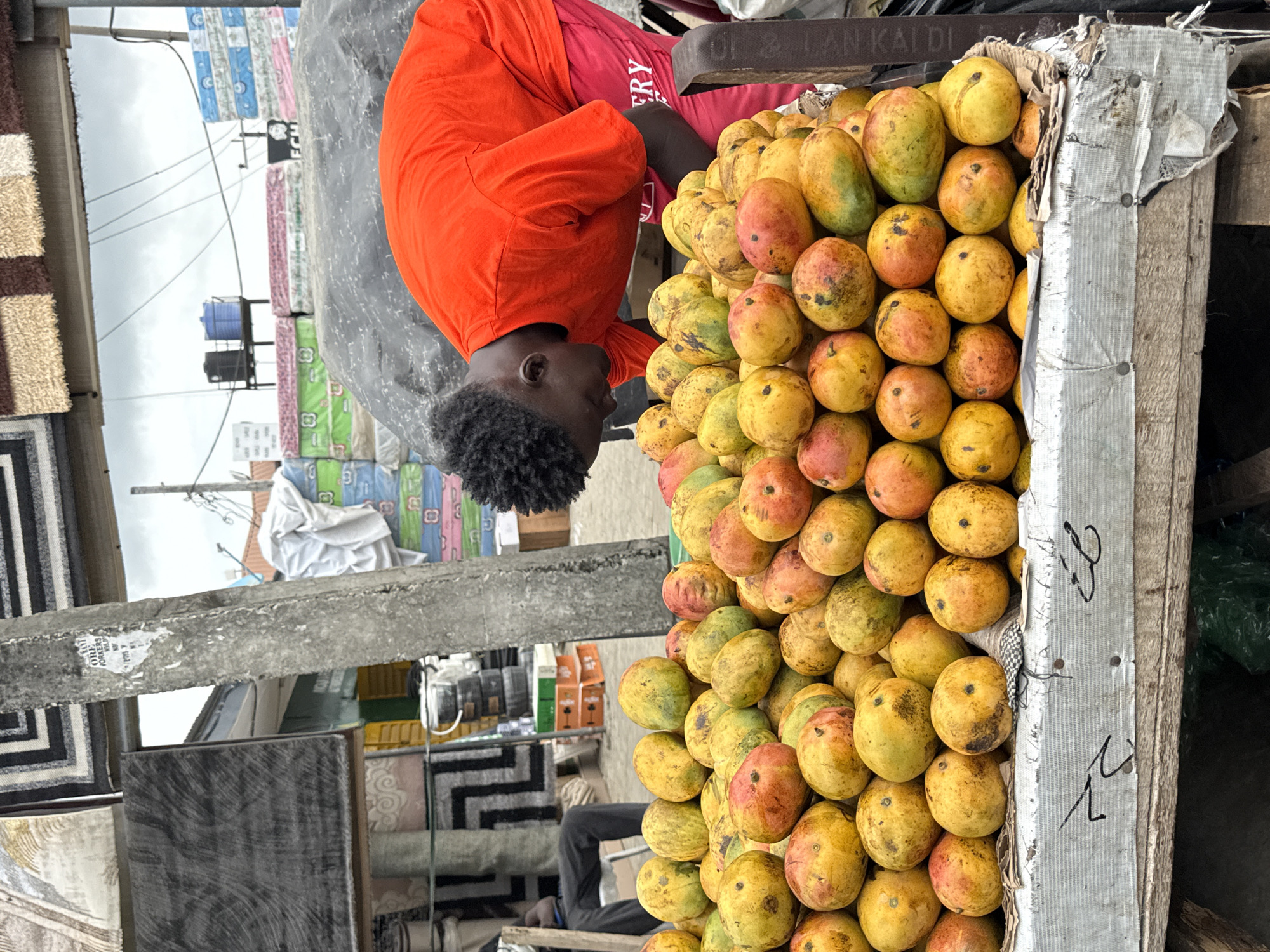 A man in an orange shirt sits behind a stack of mangoes, likely selling them. The scene takes place outdoors, possibly in a market. There are carpets and mattresses in the background, suggesting a bustling commercial area. The location appears to be Lekki, Nigeria, a city where street vending is common.