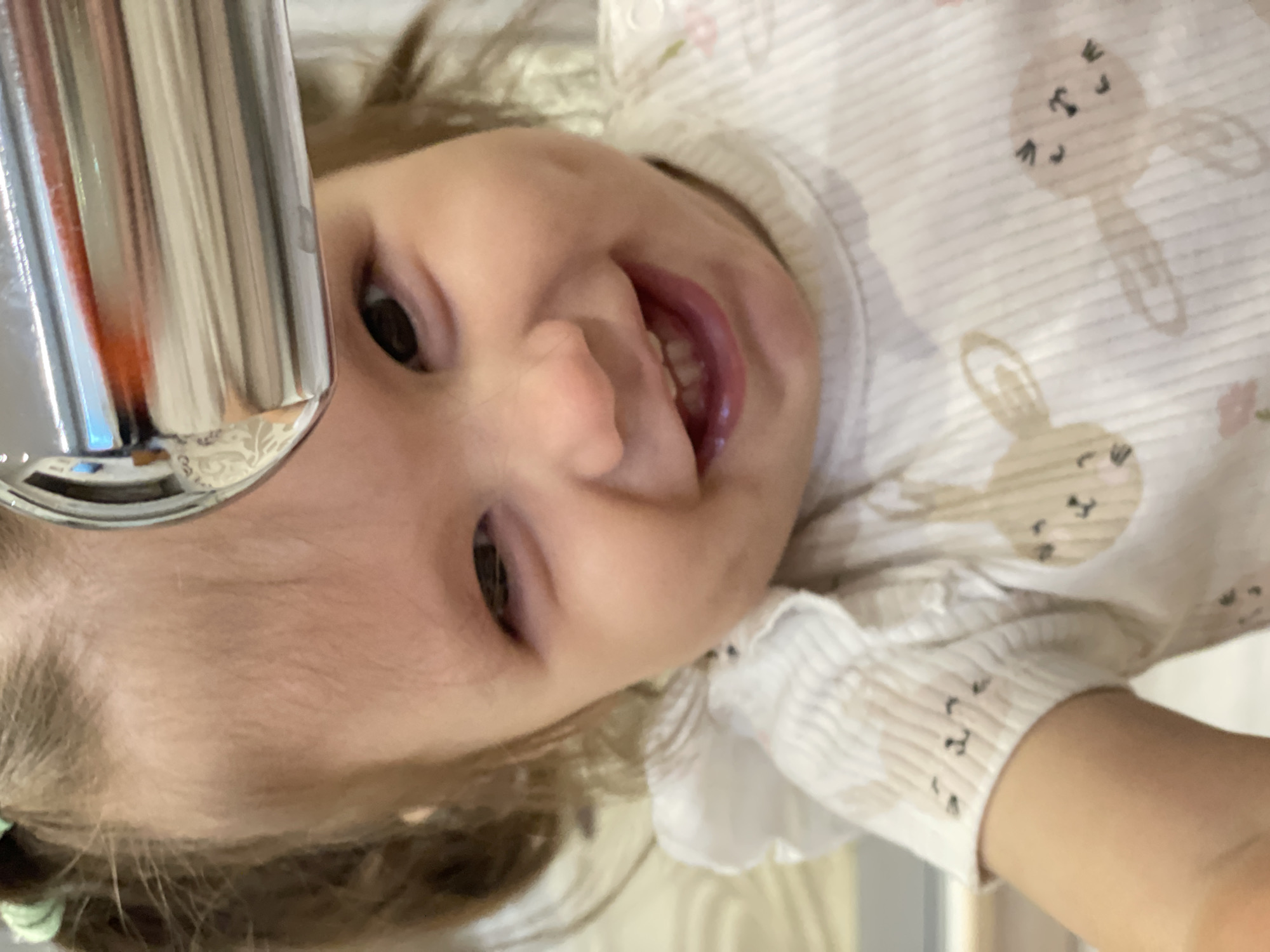 A young child, presumed to be in Izhevsk, Russia, based on the prompt, is smiling at the camera. The child is wearing a light-colored, patterned outfit with what appears to be bunny designs. A metallic object, possibly a faucet, is visible in the upper left corner of the image. The child's face is the primary focus, and their expression suggests happiness.