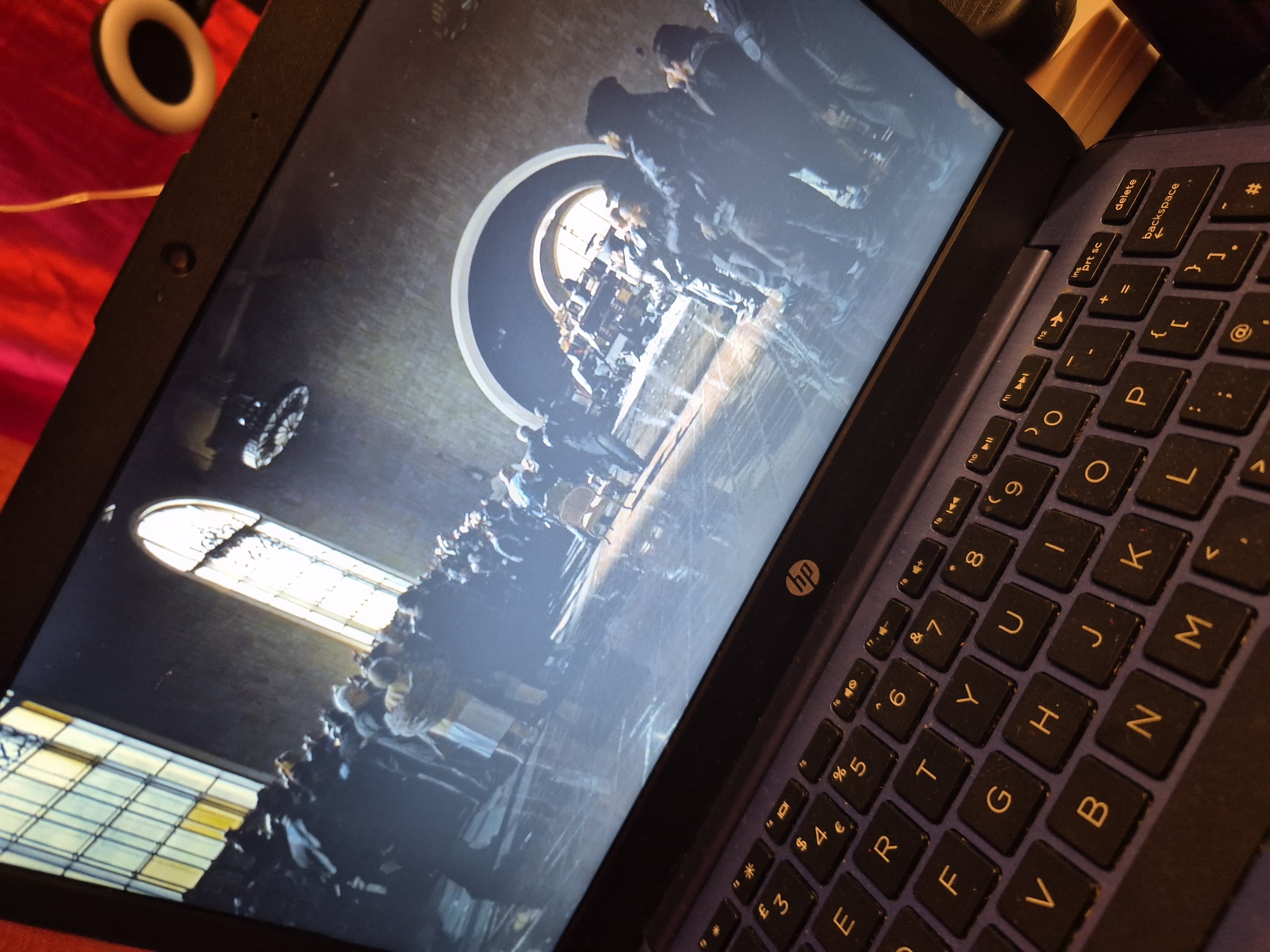 The image shows a laptop screen displaying a scene with many people gathered indoors. The room has tall, arched windows with stained glass. In the foreground, a portion of a keyboard is visible, with keys labeled with letters and numbers. The overall scene on the screen appears to be from a movie or TV show, possibly a historical drama or a period piece. The lighting on the screen is dim, with light coming from the windows and some overhead sources. The image does not provide any information to determine the location in the city of Sandwell, United Kingdom.