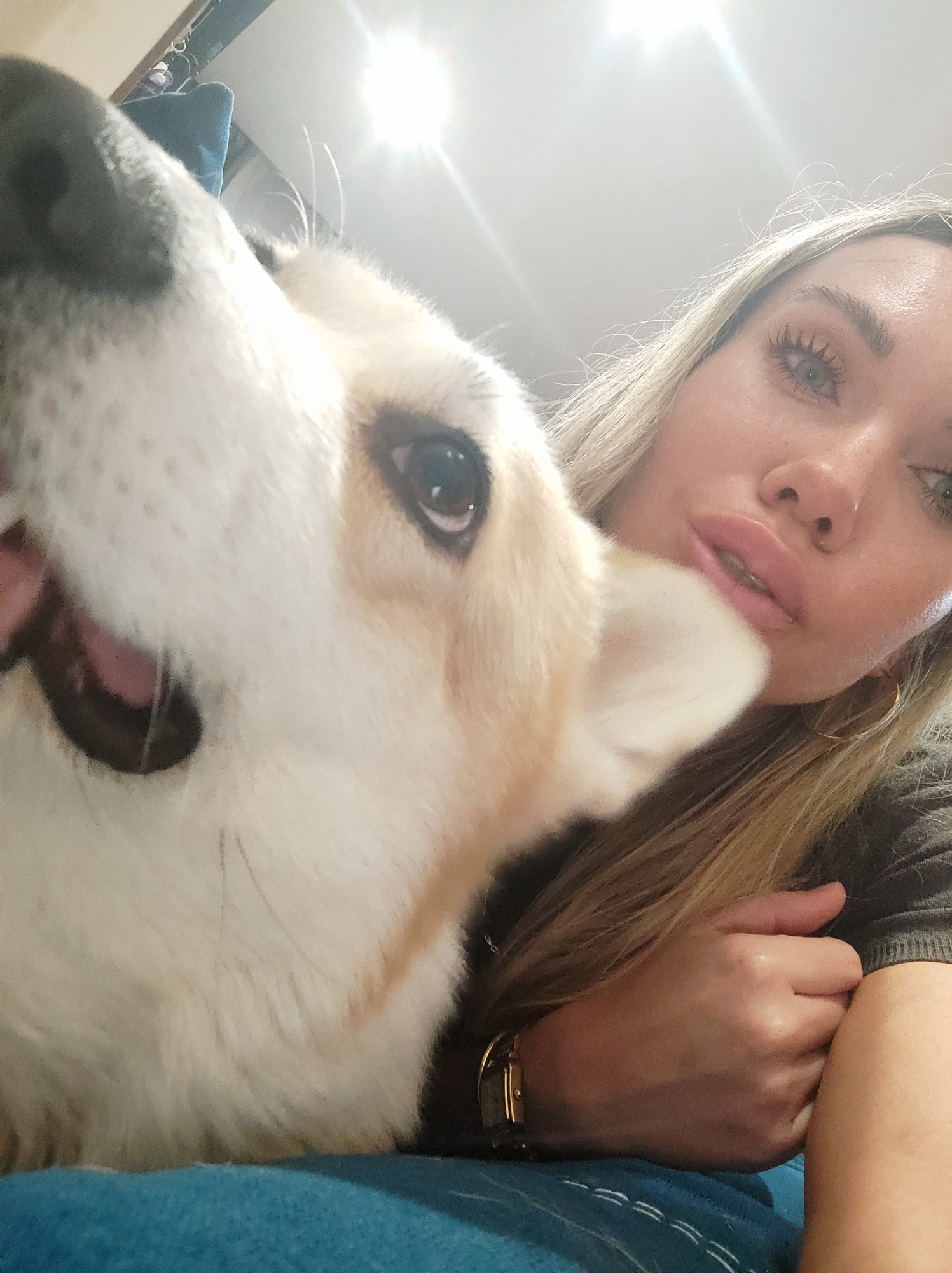 A woman with long, blonde hair and a gold hoop earring is lying on a blue surface, possibly a bed or couch, next to a corgi. The corgi's face is very close to the camera, with its nose and open mouth dominating the left side of the image. The woman's face is visible to the right, partially obscured by the dog. Her hand, adorned with a gold wristwatch, rests on the blue surface. The environment appears to be indoors, with a bright light source overhead. There is no information suggesting this image was taken in Votkinsk, Russia.