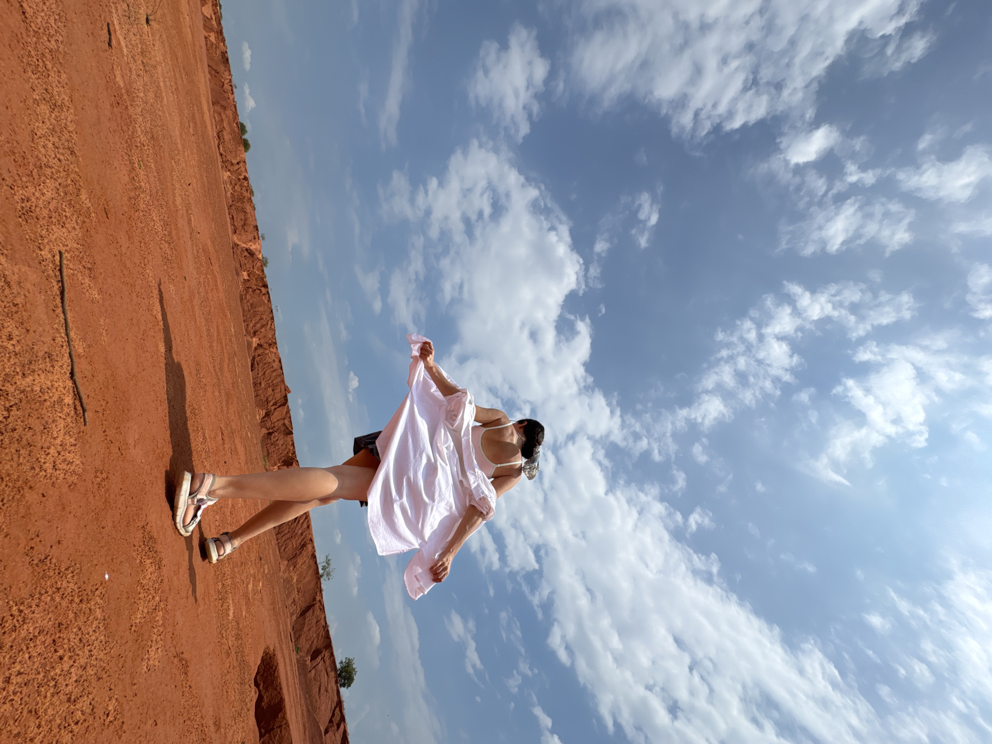 A low-angle shot shows a person from the back, wearing a white dress and sandals, standing on a red, sandy terrain. The person is holding the sides of their dress, allowing it to flare out, and is looking away from the camera. The background features a vast expanse of blue sky with scattered white clouds, and a distant red cliff formation. The scene appears to be set in a desert-like environment, consistent with the sand dunes of Mui Ne, Vietnam.