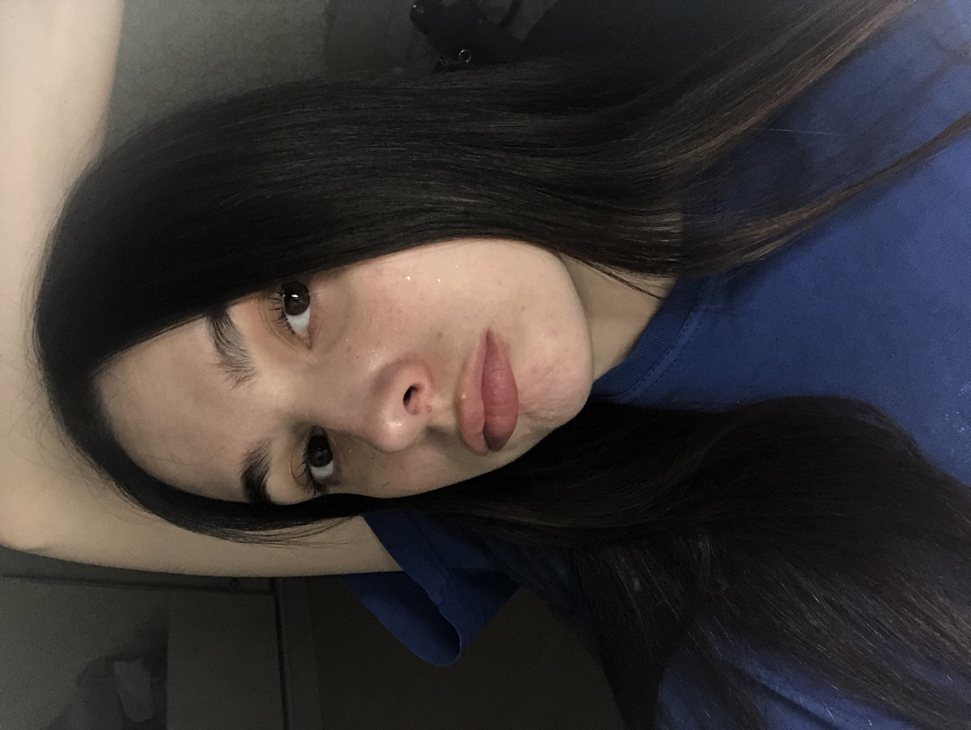 A young woman lies down wearing a blue shirt. Her long, dark hair spreads around her head. She has light skin, brown eyes, and appears to be wearing some glitter on her face. She is looking slightly upwards and to her left. The background appears to be a wall or a piece of furniture. There is no evidence to suggest this image was taken in Noviy, Russia.