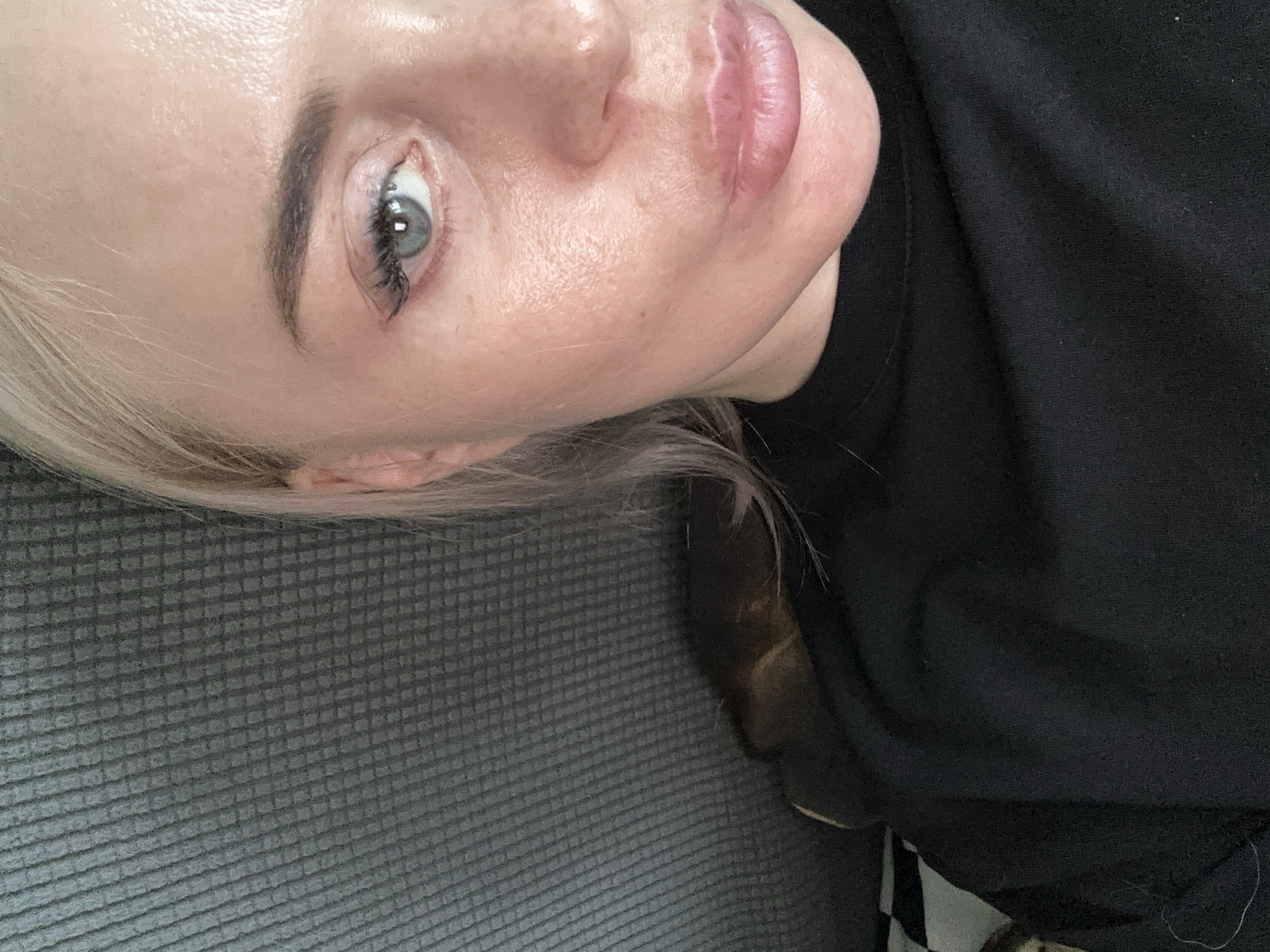 The image is a close-up, partial portrait of a young woman with blonde hair, wearing a black top, who is lying down on a textured gray surface, possibly a sofa. Her face is turned towards the camera, with her left eye visible. The lighting appears to be natural and soft, highlighting the sheen of her skin. The background is out of focus, with only hints of what might be a dark object or another person partially visible. The composition is casual, suggesting an indoor, relaxed setting. There are no specific details about a city or location.