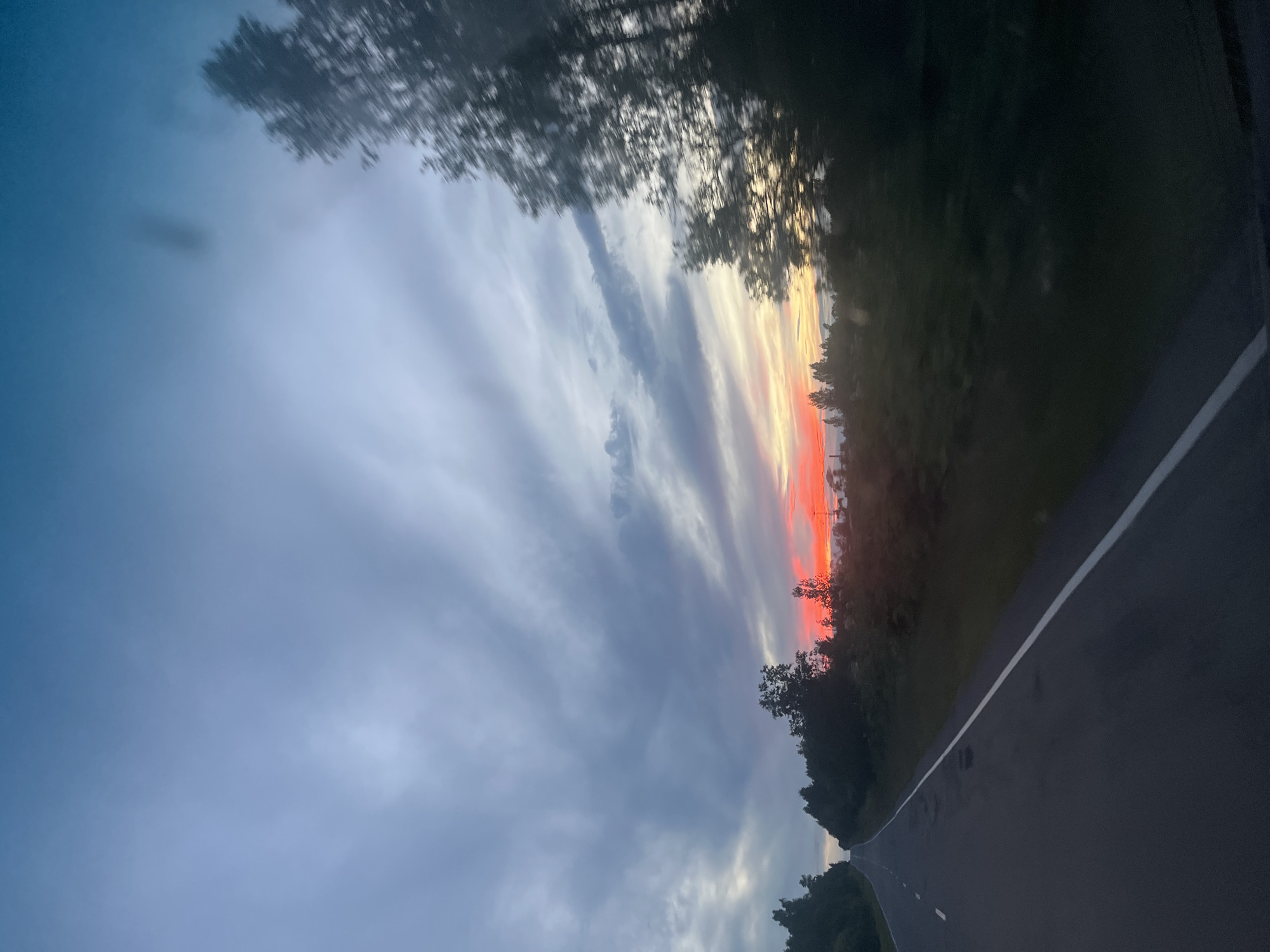The image depicts a dramatic sunset over a rural landscape. The sky is filled with elongated clouds painted in hues of deep grey, soft blues, and vibrant oranges and reds near the horizon, indicating the sun is low in the sky. The warm colors suggest it is either sunrise or sunset, likely sunset given the deepening shadows. A paved road, marked with a white line, recedes into the distance, curving slightly to the right. Trees and dense foliage line the edge of the road, creating a dark silhouette against the colorful sky. The overall mood is serene and awe-inspiring, with the natural beauty of the sunset dominating the scene. There is no visible text in the image. The location context is Серебрянский, Russia.