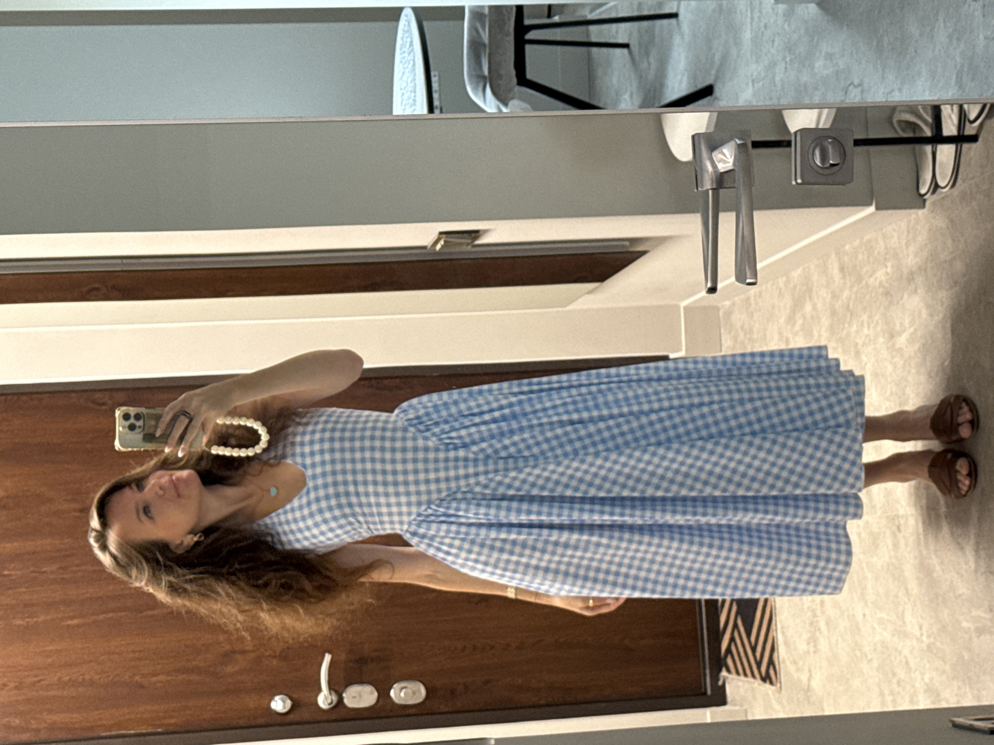 A woman with long, brown, wavy hair is standing in front of a wooden door and a mirror. She is wearing a blue and white checkered dress with a V-neckline and a full skirt. She is holding a smartphone in her right hand, taking a selfie. Around her neck, she is wearing a pearl necklace. On her left wrist, she has a gold bracelet. On her feet, she is wearing brown slide sandals. The floor is tiled with a light-colored marble pattern. In the reflection of the mirror, a small round table with a white tablecloth and a chair with a grey cushion are visible. The environment appears to be an indoor residential space.