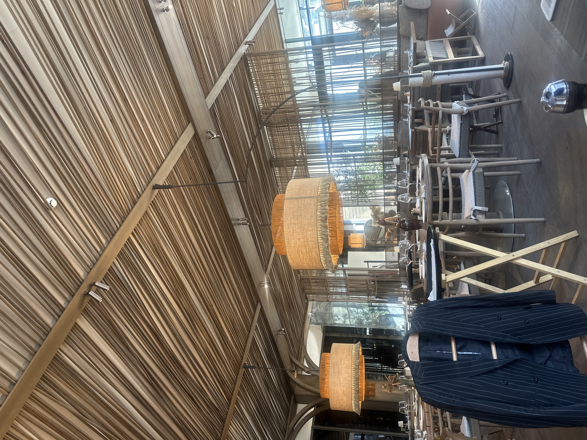 This image depicts the interior of a spacious, contemporary dining establishment or restaurant, likely situated in Kozyn, Ukraine. The scene is brightly lit by natural light, indicating it is daytime.
The most striking architectural feature is the high, vaulted ceiling, meticulously constructed from numerous thin, parallel wooden slats. These slats run diagonally across the ceiling, supported by thicker wooden beams. Several discreet recessed lights are integrated into the ceiling structure, and a small white circular object, possibly a smoke detector or sensor, is also visible.
Two prominent, large cylindrical pendant lights, each with a textured fabric shade in a natural, light brown hue, hang from the ceiling, adding a warm ambiance.
Below these lights, the main dining area is furnished with multiple tables and chairs. The chairs are crafted from light-colored wood, some featuring woven or upholstered seat cushions and backrests, often accented with rope details. Many of the tables are partially set with glassware and white plates, suggesting readiness for diners.
Large windows dominate one side of the room, allowing ample natural light to stream in and offering a glimpse of an outdoor green space or garden. Reflections of the interior elements are visible in the glass panes.
In the foreground, on the right side of the frame, a dark pinstriped blazer or jacket is draped over what appears to be a wooden chair or a display stand, adding a touch of human presence to the otherwise empty space. No people are directly visible, and no specific activity is overtly taking place, but the setup clearly indicates a dining or social gathering area. There is no visible text in the image.