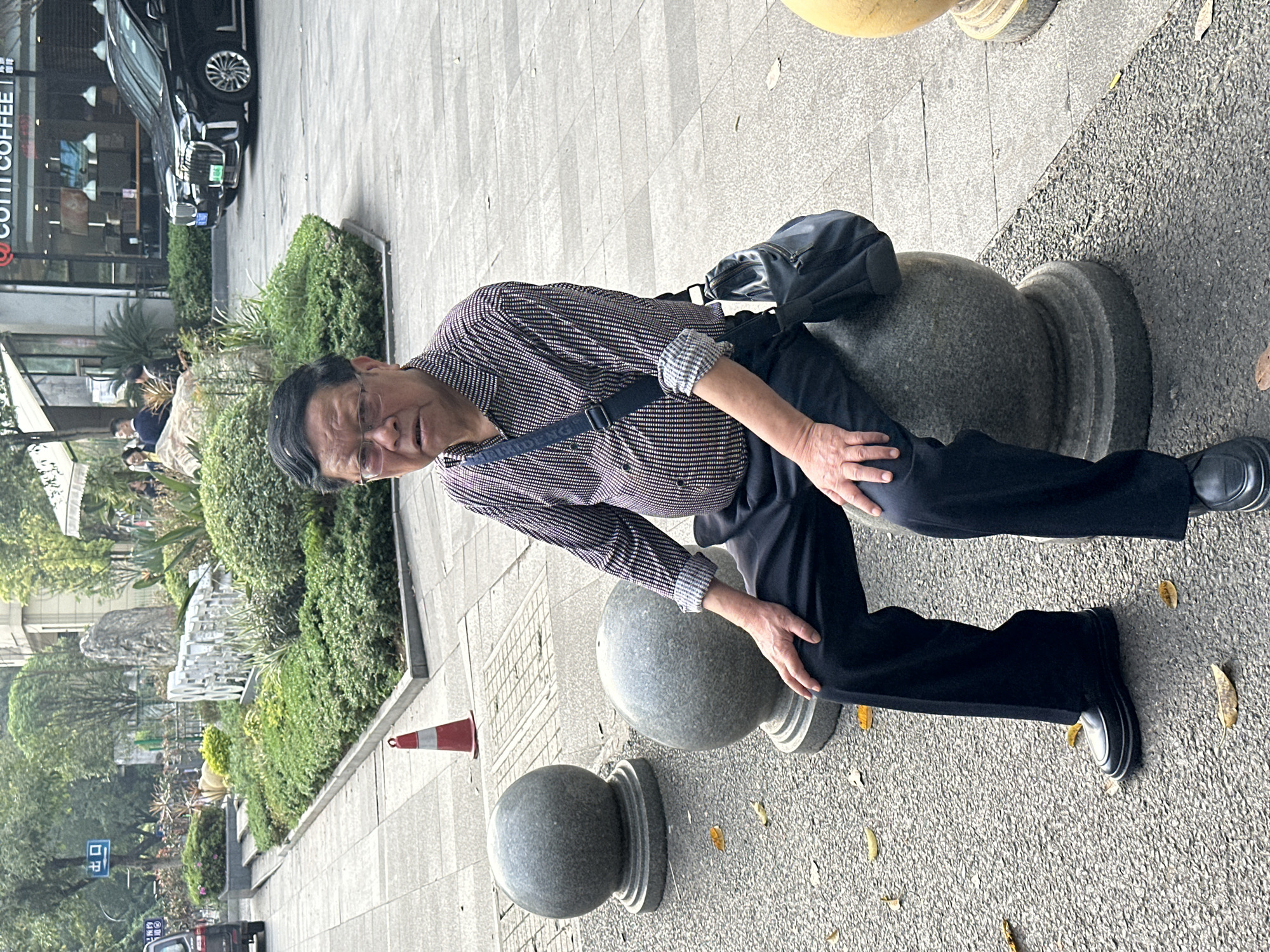 A man is seated on a series of stone bollards in an urban area, likely in Yuzhong District, China. He is wearing a long-sleeved, dark-patterned shirt, black trousers, and black shoes. A messenger bag is slung across his body. He appears to be resting, with his hands placed on his knees. The bollards are gray and spherical, each mounted on a short, square base. A traffic cone is visible on the paved sidewalk in the background, along with greenery, trees, and a building with a "COSTA COFFEE" sign. A car is parked in front of the building. The pavement is gray and sprinkled with fallen leaves.