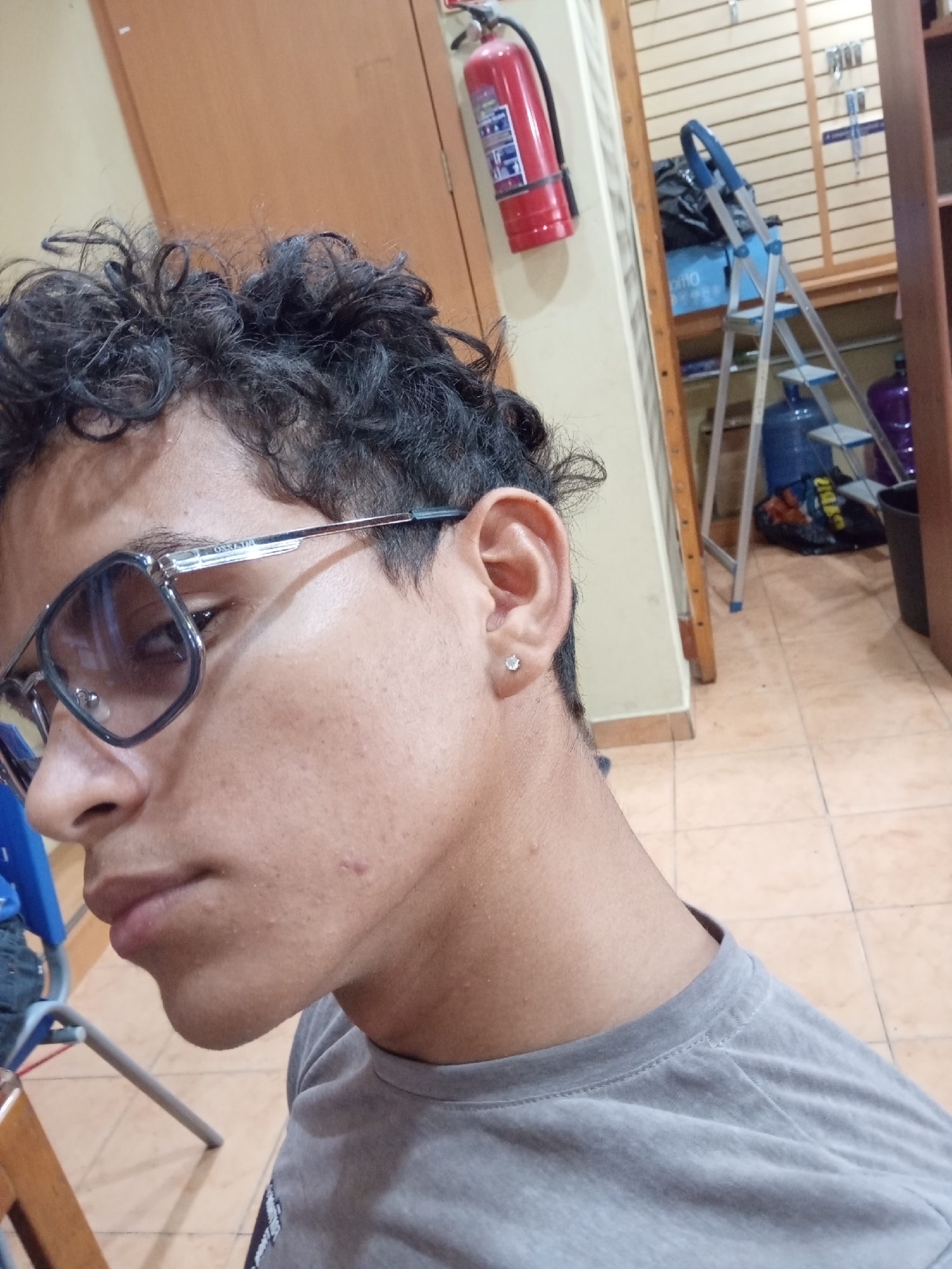 A close-up shot shows a young man with curly black hair wearing silver-framed glasses and a gray t-shirt. He has a small stud earring in his left ear.
In the blurred background, a red fire extinguisher hangs on a light-colored wall near a mirror reflecting a stepladder, blue water jugs, and shelving. The floor is tiled in a light tan color.
The location (Juan Griego, Venezuela) is not directly apparent from the visual information.