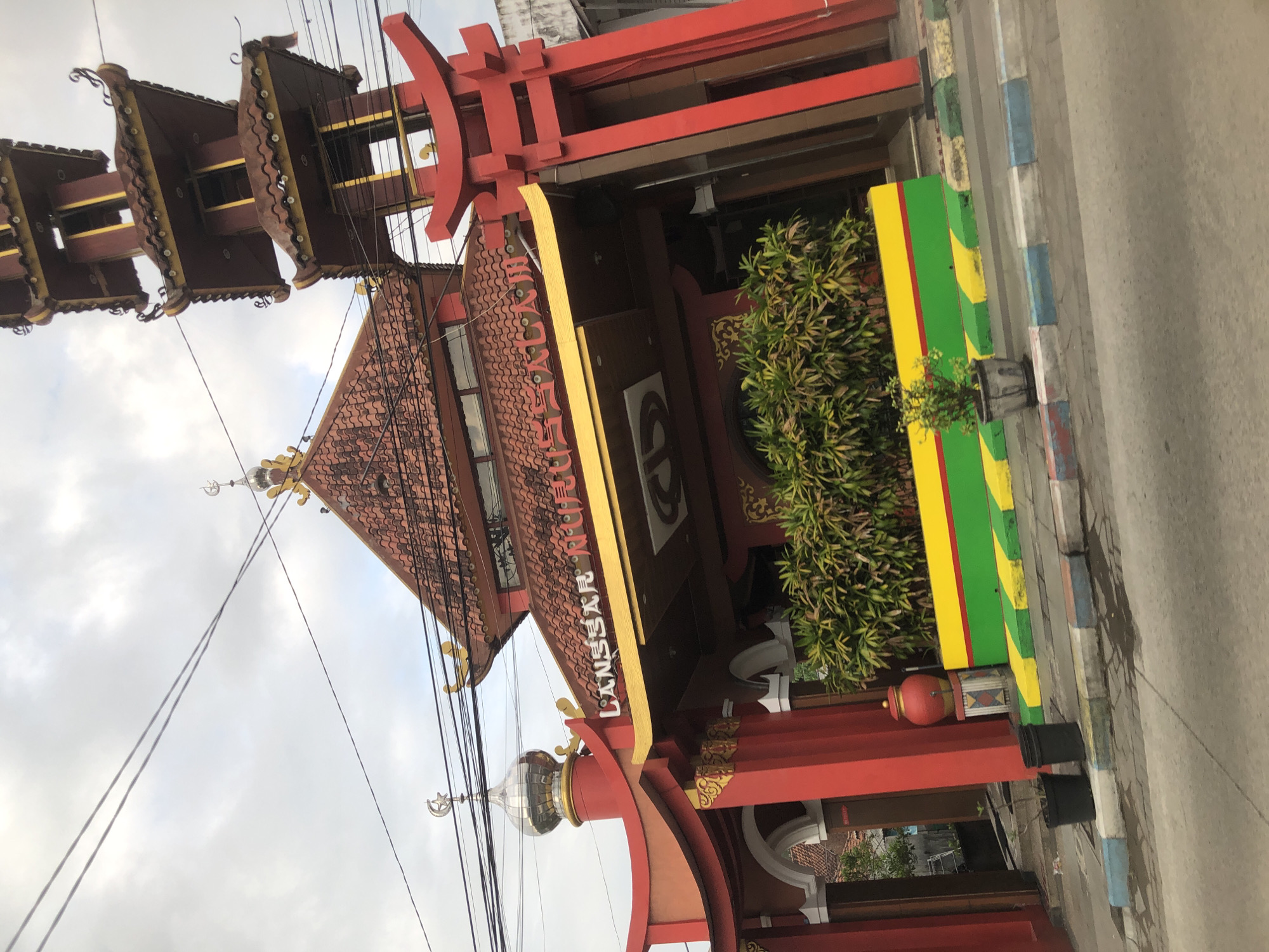 The image shows a red building with a pagoda-style tower, located in Boro, Indonesia. The building has a tiled roof and is adorned with Islamic crescent moon symbols and what appear to be Torii gates, suggesting a blend of architectural styles. The entrance features a yellow awning with the white text "LANGGAR MUSHOLA" visible. In front of the building, there is a low wall painted with horizontal stripes of yellow, green, and red. Several potted plants are visible, including two black planters near the entrance and a small plant in a decorative pot in front of the striped wall. The scene is viewed from a street level, with power lines crisscrossing the sky above.