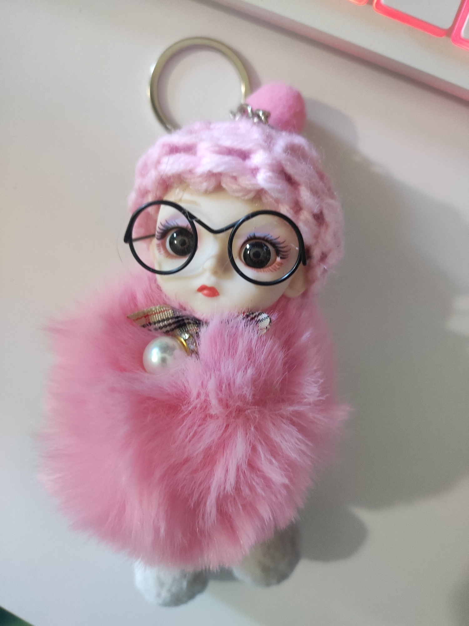 The image displays a close-up, high-angle shot of a pink keychain charm shaped like a doll. The doll has fair skin, large brown eyes with prominent eyelashes, and red lips. It wears a knitted pink hat with a small pink pom-pom and a silver charm on top. Large, round, black-rimmed glasses are perched on the doll's nose. The doll is enveloped in a fluffy pink fur body, and a single, iridescent pearl hangs from its neck area, attached to a patterned ribbon. The background is a plain, light-colored surface, likely a desk or table, with a hint of a pink keyboard visible at the top right edge of the frame. There is a soft shadow cast to the right of the doll. The scene does not depict any specific location in Cherkasy, Ukraine, or any identifiable landmarks.