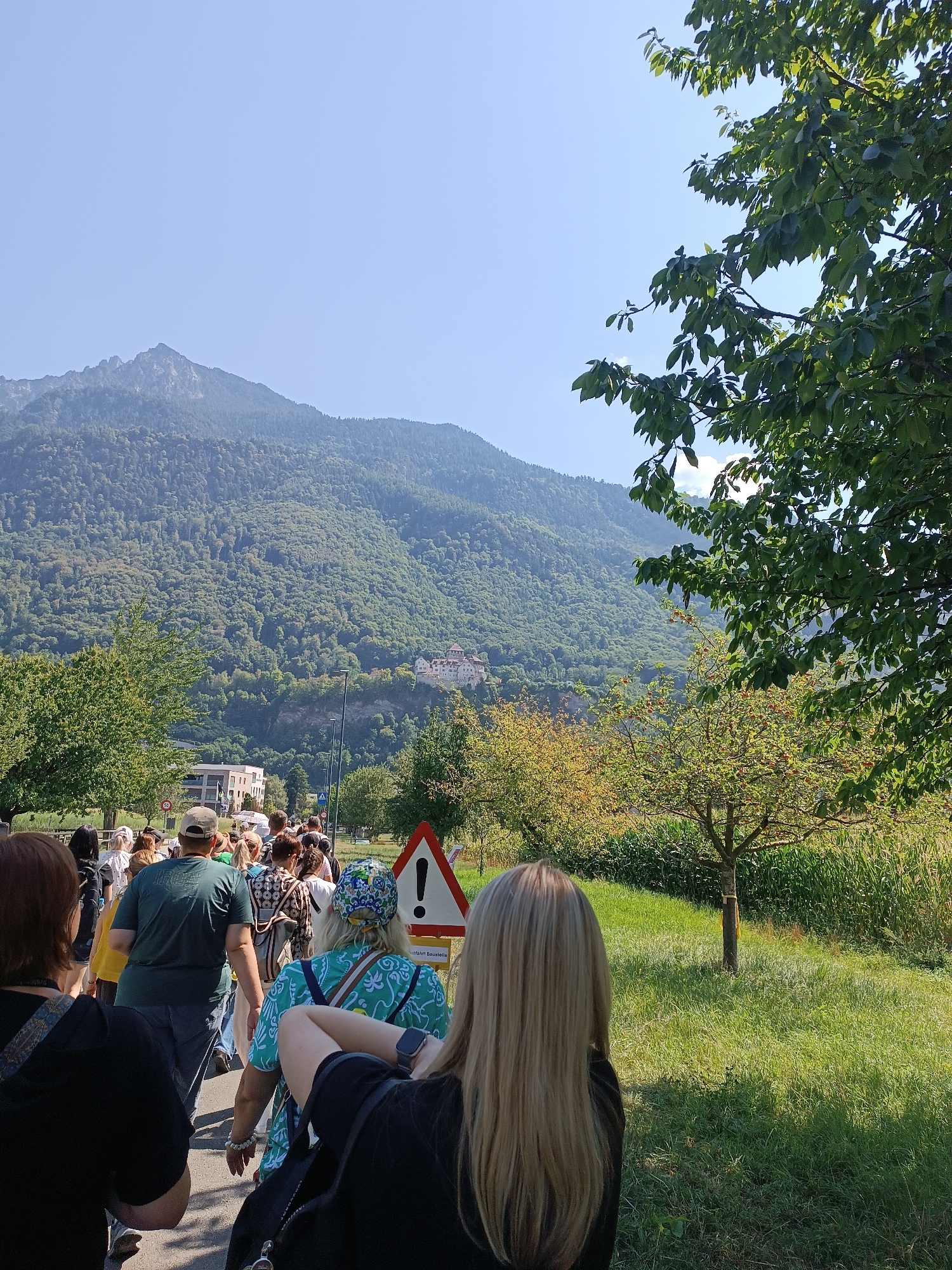 The image shows a group of people walking down a paved path on a sunny day. They are walking away from the camera, towards a mountain range covered in lush green trees. In the distance, perched on a hillside, is a large, white building complex with a red-tiled roof. To the right of the path is a grassy field with a small tree. Several traffic signs are visible along the path, including a triangular warning sign with an exclamation mark. The sky is clear and blue. The overall impression is of a scenic outdoor excursion in a mountainous region, possibly a hiking trail or a route leading to a tourist attraction.