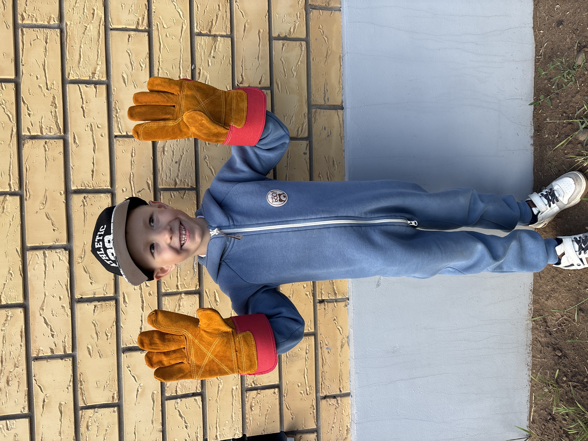 A young child, likely a boy, stands smiling brightly for the camera in an outdoor setting in Chaikovsky, Russia. He is dressed in a blue, full-body jumpsuit with a white zipper down the front and white stripes on the side seams. A circular logo featuring a cartoon animal wearing a cap is visible on the left chest of the jumpsuit. His hands are raised and covered in oversized orange work gloves with red cuffs, suggesting play or an activity. He wears a black and beige baseball cap with the word "ATHLETIC" partially visible across the front. White sneakers with black laces and Velcro straps are on his feet.
The background to the left features a textured light brown brick wall, indicating the exterior of a building. To the right, there is a smooth, light grey concrete path or slab, and beyond that, a strip of dirt with sparse green grass. The lighting is bright and even, characteristic of daytime. The child's wide smile and posture convey a sense of happiness and playfulness.