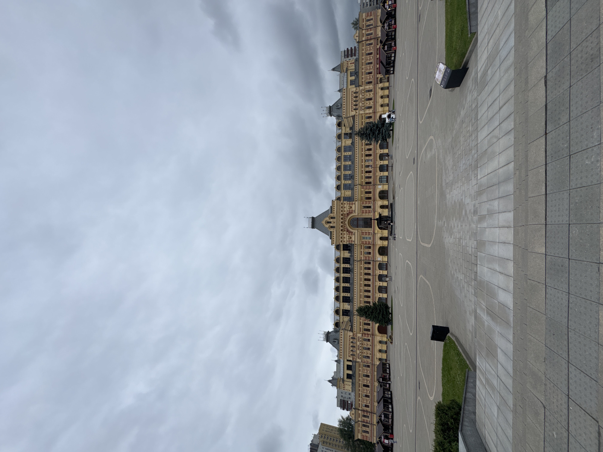 The image shows a wide, paved plaza in front of a grand, yellow building with ornate architecture and numerous windows. The building appears to be historic, with multiple stories and a complex roofline featuring spires and decorative elements. Several small, dark structures, possibly market stalls or kiosks, are lined up along the side of the building. A few evergreen trees are planted in the plaza. The ground is paved with large, rectangular stones, and there are white, curved lines painted on a section of the plaza, possibly indicating a designated area. A black information stand is visible to the right of the building. The sky is overcast with heavy, grey clouds, suggesting a dull or possibly rainy day. The orientation of the image is vertical, with the building running from the bottom to the top of the frame. The overall scene depicts an urban public space, likely a historical square or marketplace, under a somber sky.