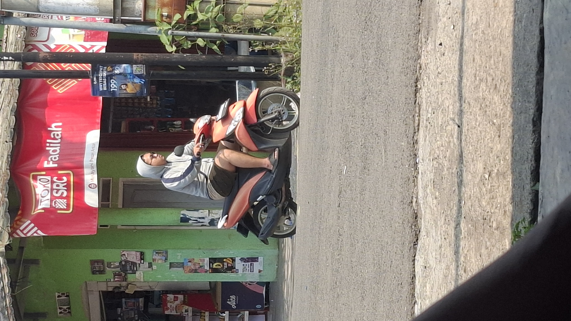A young Indonesian male wearing a grey hooded jacket and dark shorts is riding a red and orange scooter on an asphalt road in front of a green building. The building has a red banner overhead that reads "TOKO SRC Fadilah" and "Melayani Pesan Antar." A pole with a flyer advertising a phone plan is visible to the right of the building. The road is paved and appears to be a street in a populated area, possibly a commercial district given the building's storefront. The weather appears to be sunny.