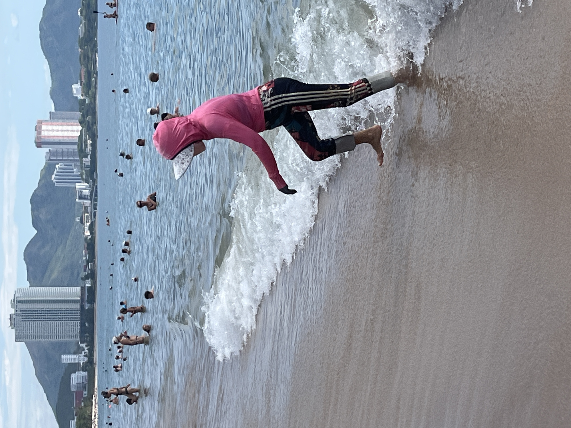 This image captures a beach scene in Nha Trang, Vietnam, with the city skyline in the background. In the foreground, a person wearing a pink hooded rash guard and patterned leggings is wading through shallow water near the shore. The person is barefoot on their right foot and has a gray cover on their left leg. Waves are breaking around their legs. In the middle ground and background, numerous people are visible in the water, some swimming and others standing. The water is a clear blue, and the sky is partly cloudy. The buildings in the distance suggest an urban environment bordering the beach.