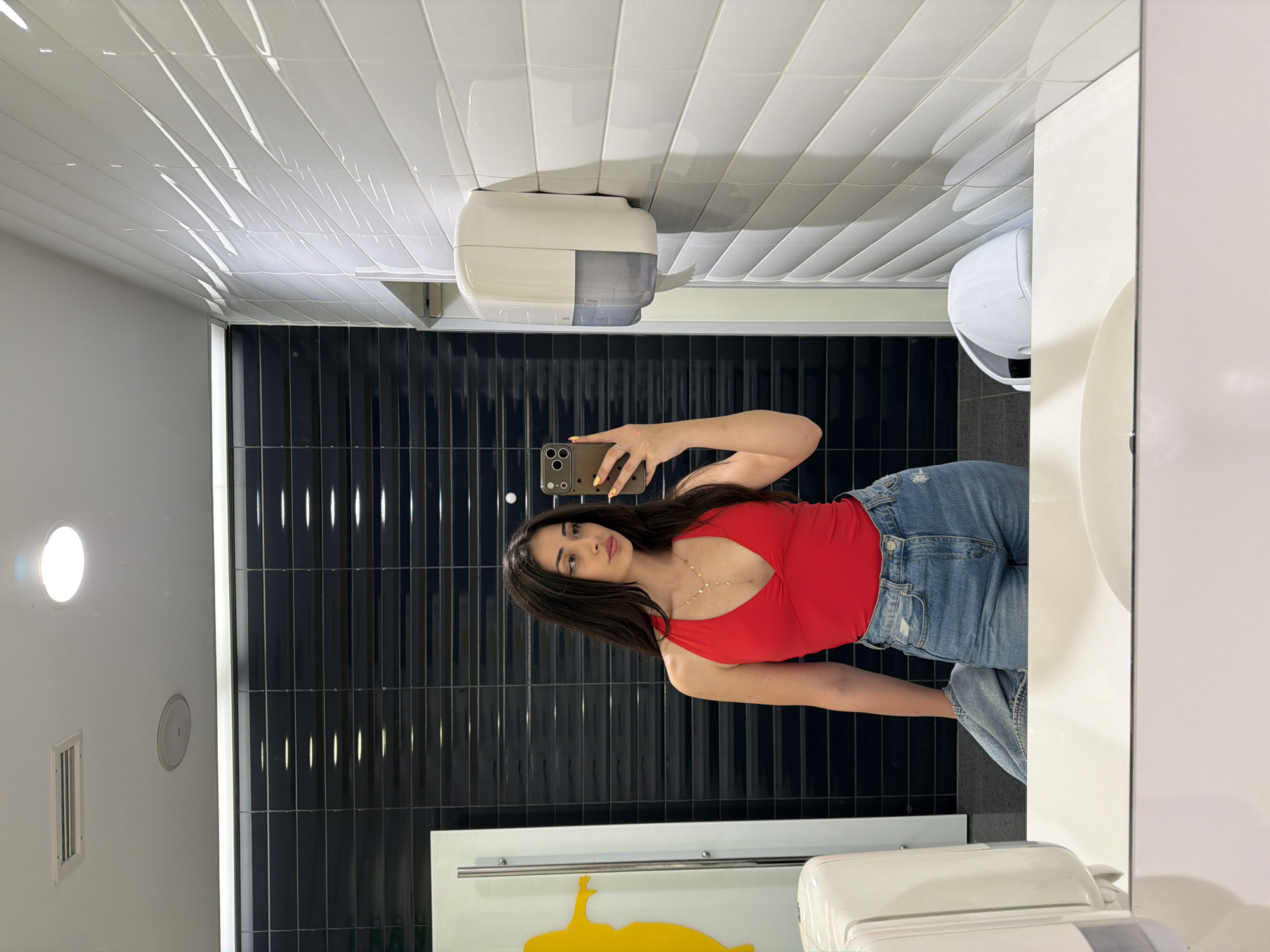 A young woman with long dark hair is taking a selfie in a public restroom. She is wearing a red halter top and denim shorts. She is holding a smartphone in her right hand, and a denim jacket is draped over her left arm. The restroom has black tiled walls in the background, and white tiled walls on the right. There are two dispensers mounted on the walls, one white and the other with a clear reservoir. A sink is visible in the foreground, reflected in the mirror. The lighting is bright, suggesting overhead fluorescent or LED lights. The scene appears to be in a modern, public facility.
