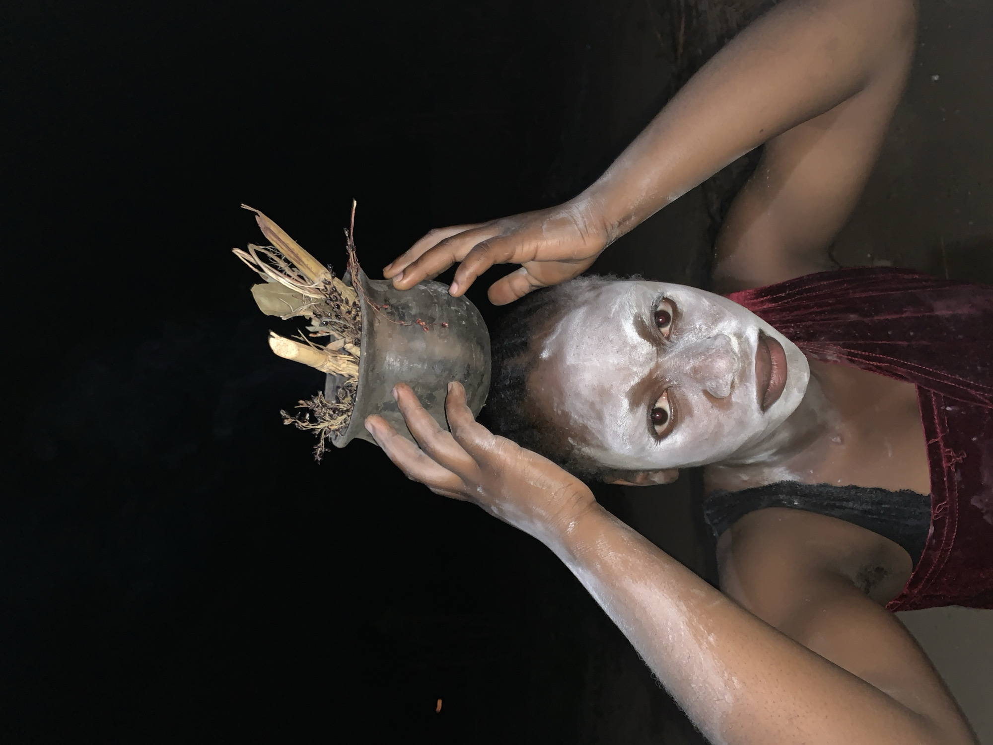 A woman with white face paint is holding a pot with dry plants on her head. She is wearing a maroon top with black lace trim. The background is black, possibly a dark room. The exact location in Kukum, Nigeria cannot be determined from this image.