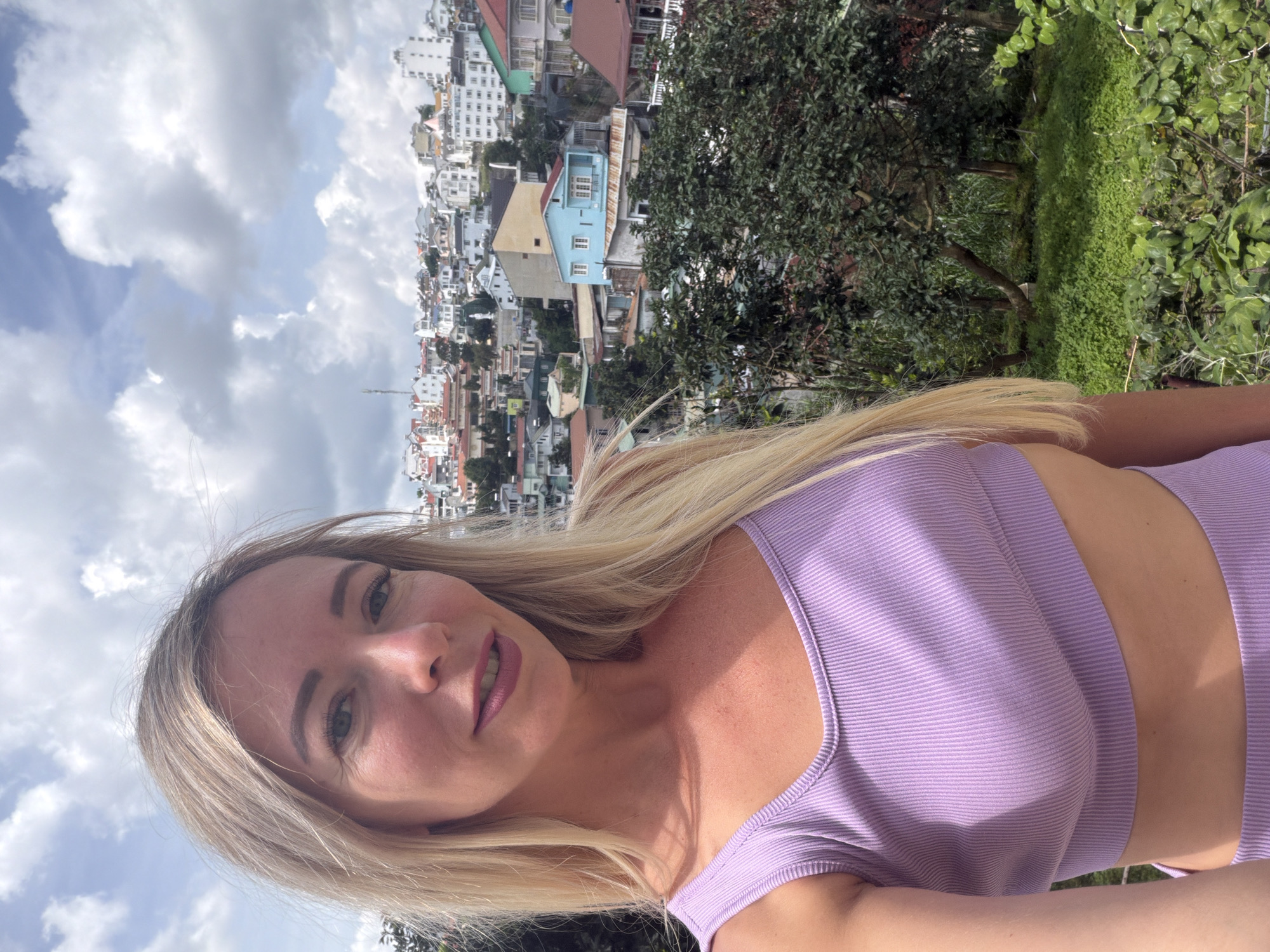 A woman with blonde hair, wearing a light purple crop top, takes a selfie outdoors. Behind her, a city with numerous buildings of various colors and sizes is visible on a hillside under a cloudy sky. Lush green trees and foliage are in the foreground and mid-ground, with some buildings peeking through the trees. The scene appears to be in a hilly urban area, consistent with the topography of Da Lat, Vietnam.