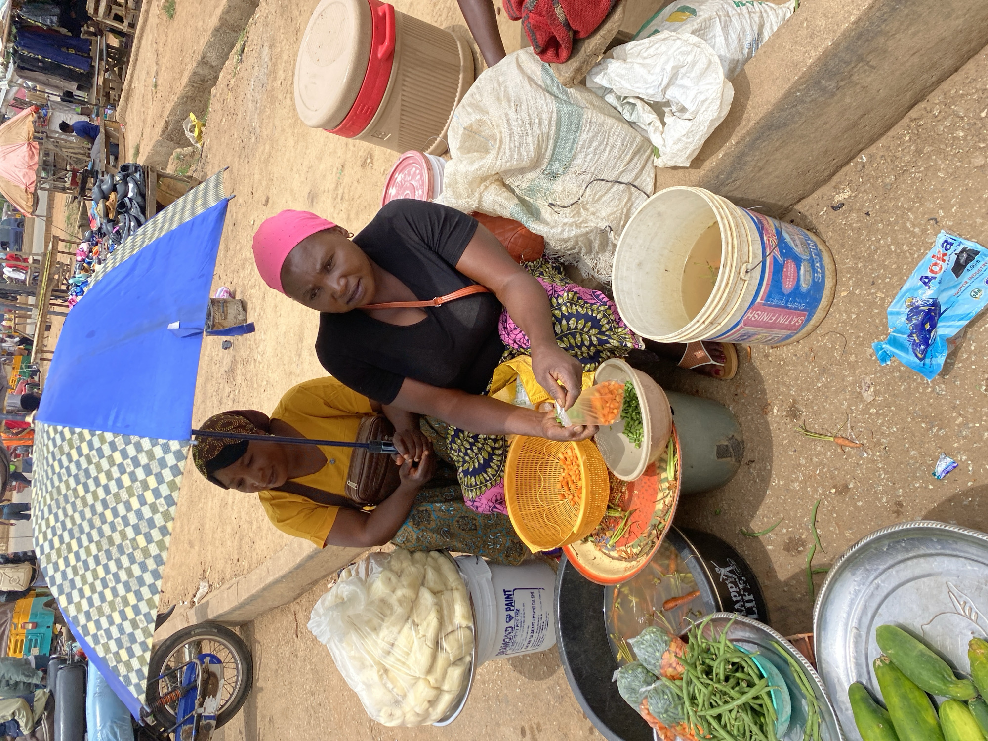 The image depicts an outdoor market scene in Bukuru, Nigeria. Two women are the central figures, seated and attending to produce. The woman on the left wears a yellow shirt and a patterned headscarf, while the woman on the right wears a black shirt and a pink headscarf. They are positioned near a large blue and white checkered umbrella, which provides shade.
In front of the women, various food items and containers are arranged. A large plastic bucket, labeled "DIAMOND PAINT," holds a substantial quantity of pale, oblong food items wrapped in plastic. To its left, a dark basin contains water and various vegetables, including green beans and what appear to be carrots. A yellow plastic colander is filled with chopped carrots, and a smaller bowl holds chopped green beans. A woman's hand is visible, holding a plastic bag of chopped green beans. Further to the right, a large beige cooler with red accents is visible, along with several bags, one of which is white and appears to be made of woven material. A partially unwrapped blue plastic package with text and an image of a vehicle lies on the ground.
The ground is a mix of sand and dirt, with scattered debris. In the background, the market extends with wooden stalls displaying clothing and footwear, and a few other individuals are partially visible, suggesting a busy marketplace. The overall environment is a typical street market setting, with the presence of umbrellas, various containers for goods, and a sense of commercial activity.