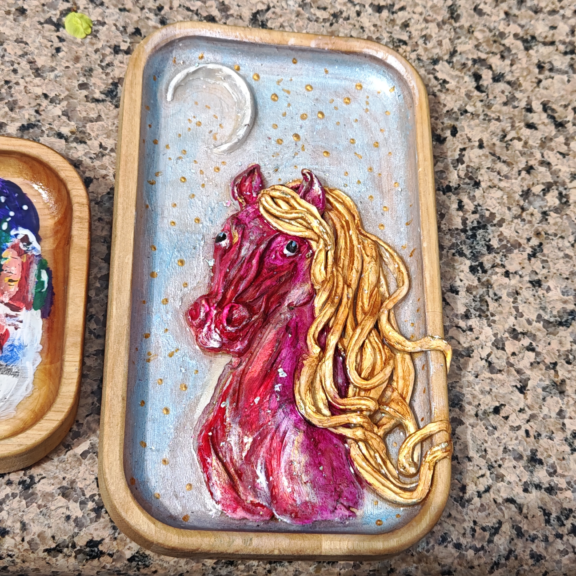 A rectangular wooden tray displays a 3D relief art piece featuring a red and pink horse with a flowing gold mane against a pale blue background with gold speckles. A white crescent moon is located in the upper left corner of the background. The tray rests on a speckled brown and beige surface. A portion of another wooden tray with artwork is visible on the left. There is no indication of Taganrog, Russia.