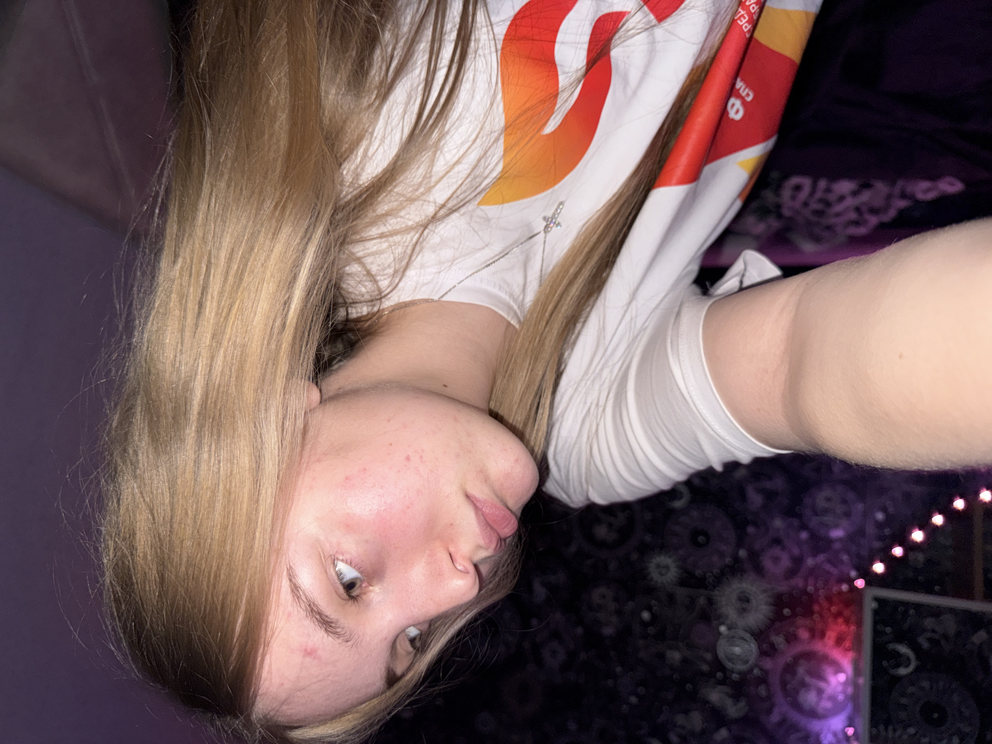The image is a close-up, low-angle, indoor selfie of a young Caucasian female with long blonde hair. She is wearing a white t-shirt with a colorful graphic design featuring red and orange flame-like shapes and some text. She also wears a silver necklace with a small cross pendant. Her head is tilted back, and she is looking upwards and to the right. Her skin appears to have some redness and minor blemishes. The background is dark and features a patterned tapestry with purple and pink lighting effects, and a string of small pink lights. The overall lighting is dim and appears to be artificial, possibly from the string lights or a nearby light source. There are no discernible architectural details or city landmarks that would indicate a specific location within a city in Russia.