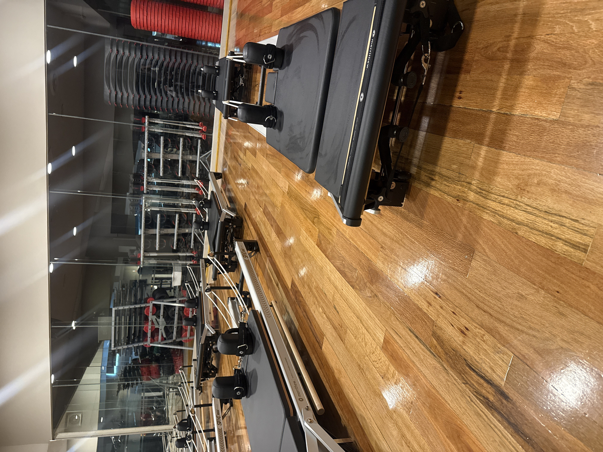 This media file captures an indoor pilates studio, likely part of a larger fitness center in Puebla, Mexico. The scene is dominated by multiple Pilates reformers arranged side-by-side on a polished wooden floor.
In the foreground, a black Pilates reformer is prominently featured, showing its padded carriage, foot bar, and various straps for exercises. The brand name "Merritthew" is visible on the side of this equipment, with additional smaller text that appears to indicate "STOTT PILATES" and "Made in Canada." Further down the line, other similar reformers are visible, with "SPX Max" identifiable on one.
To the left, a large mirrored wall reflects the interior of what appears to be a weight training or general gym area. The reflections reveal racks laden with stacked weights, some red and some black, along with other exercise equipment. Overhead lighting fixtures are also reflected in the mirror, casting bright beams.
The floor is made of highly polished, light-brown wooden planks, showing distinct grain patterns and reflecting the studio lights and equipment. There are no people present in the image, indicating the studio is empty at the moment, prepared for future use. The overall setting suggests a professional and well-maintained fitness environment. The image is captured from an angled perspective, creating a dynamic diagonal composition across the floor and equipment.