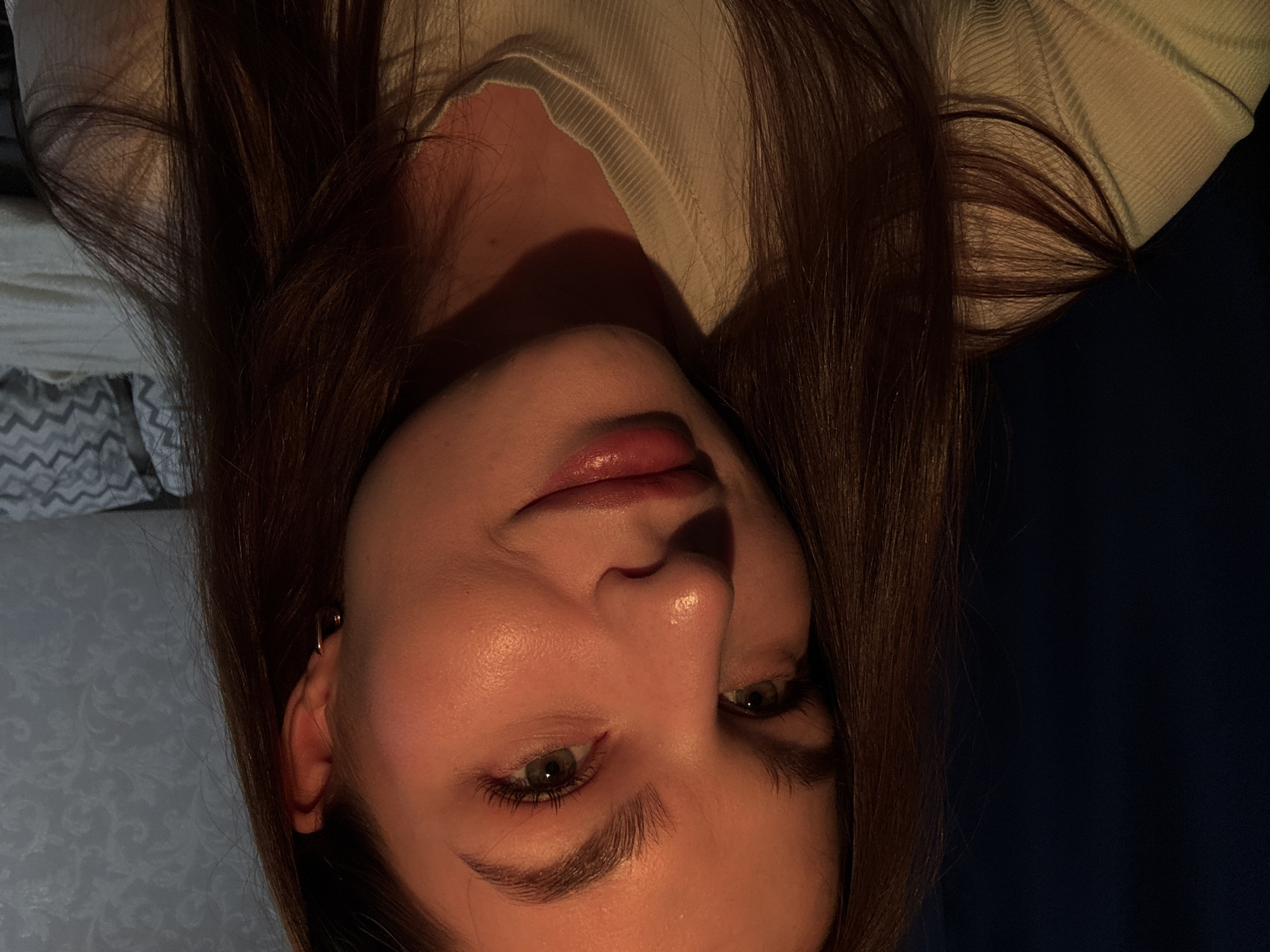 A close-up, portrait-style photograph depicts a woman with fair skin and long, brown hair. The image is rotated, presenting her face at an unusual angle. She has green eyes, defined eyebrows, and slightly parted lips with a glossy appearance. A subtle earring is visible on her left ear. She is wearing a light-colored, ribbed garment. The background consists of a dark blue or black area on one side and a patterned gray and white surface on the other. There is no information to ascertain the location as Chaikovsky, Russia.