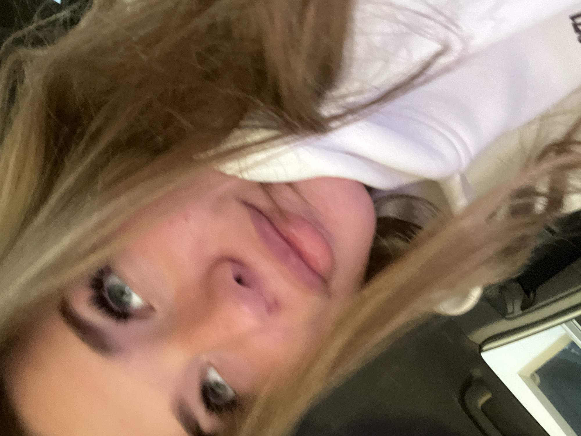 A close-up, angled portrait depicts a female individual in Budva, Montenegro. She possesses blonde to light brown hair, fair skin, and blue-green eyes, with a small, dark mark visible below her nose. The individual is looking upwards and to the left, wearing a white hoodie, which partially obscures the lower right side of her face. The background is predominantly dark and out of focus, though a blurred structural element, consistent with a window frame or a vehicle interior, is discernible in the lower left quadrant. No specific actions or explicit interactions are observed beyond posing for the photograph.