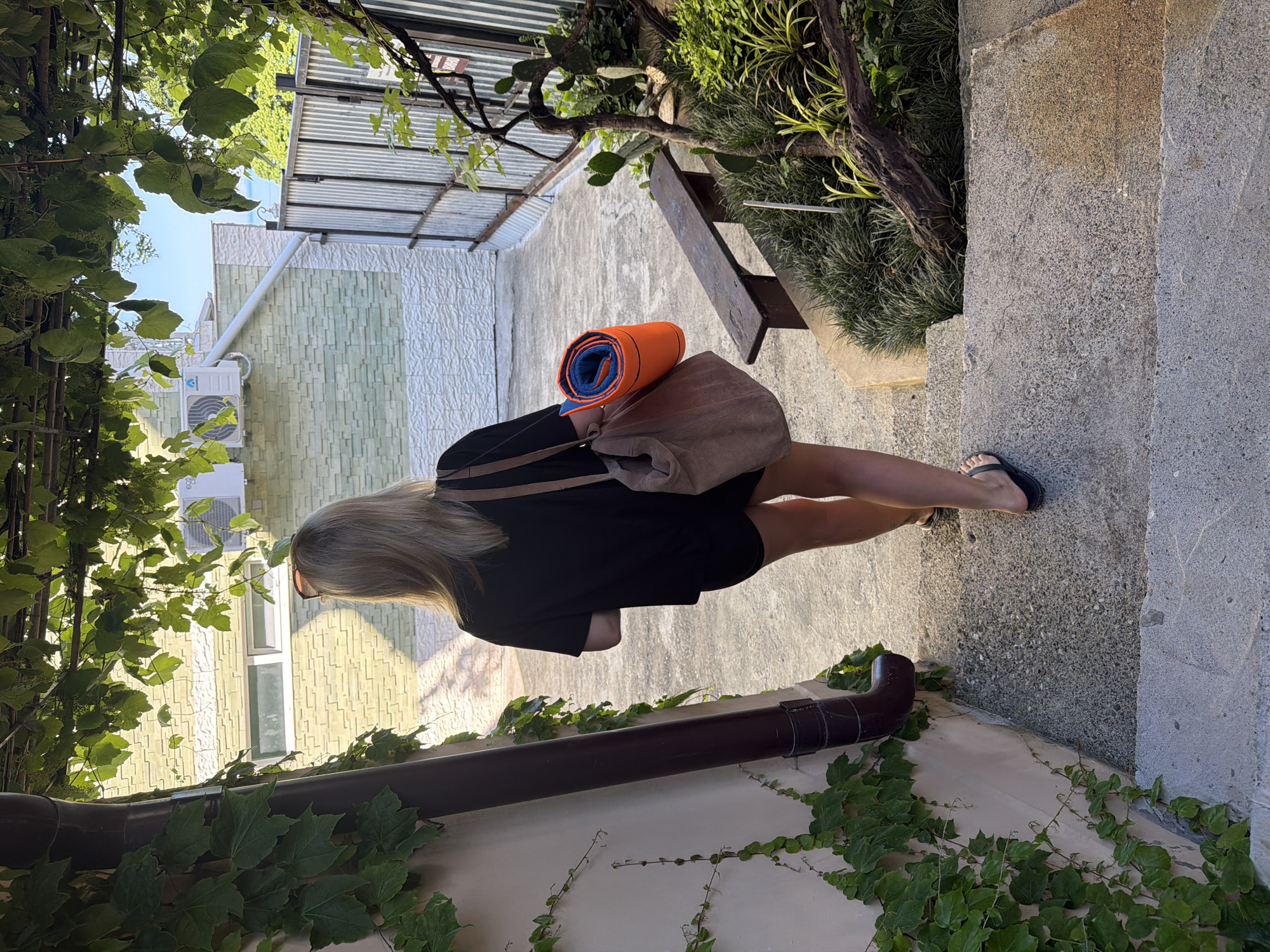 A blonde woman with shoulder-length hair, wearing a black t-shirt and black shorts, walks down a concrete staircase. She carries a brown tote bag on her left shoulder and an orange yoga mat rolled up in her right hand. The staircase is flanked by a beige wall with green ivy and a dark brown downspout on the left, and a wooden bench and lush green vegetation on the right. In the background, a building with light green and white patterned walls and an air conditioning unit is visible, along with a corrugated metal fence and a glimpse of the sea under a clear blue sky. The scene appears to be set in a courtyard or garden area, possibly in Sochi, Russia, given the presence of the sea and lush greenery.
