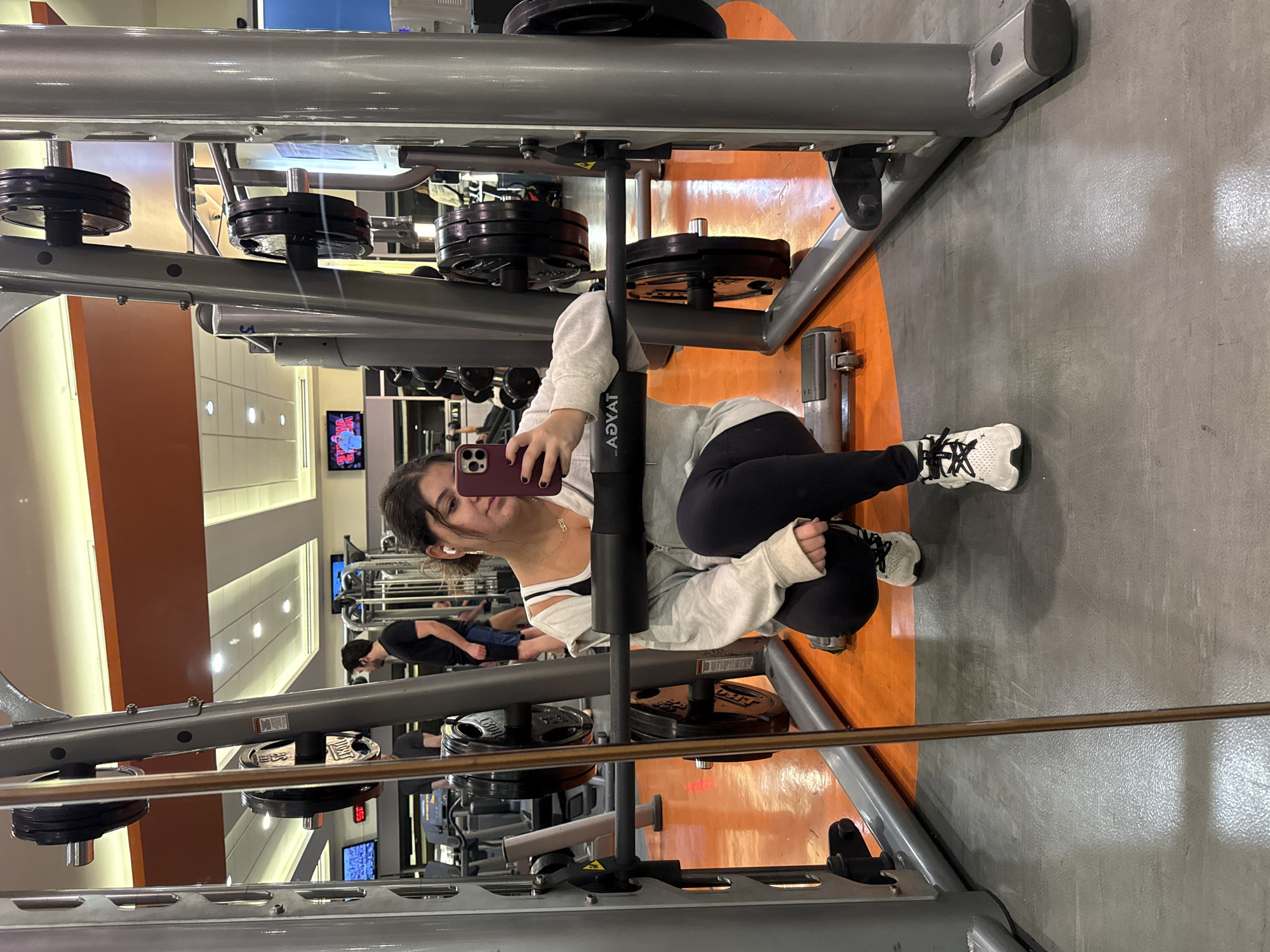 A woman in workout attire is taking a selfie while squatting inside a weight machine in a gym. She is wearing black leggings, a white top, a light grey hoodie, and white sneakers. She is holding a maroon-colored smartphone. The weight machine is gray and black with weights loaded on each side. Other people and workout equipment are visible in the background, indicating a typical gym setting. The gym has orange and gray flooring. The location is likely Mexico City, Mexico, as the make of the machine is Advat.