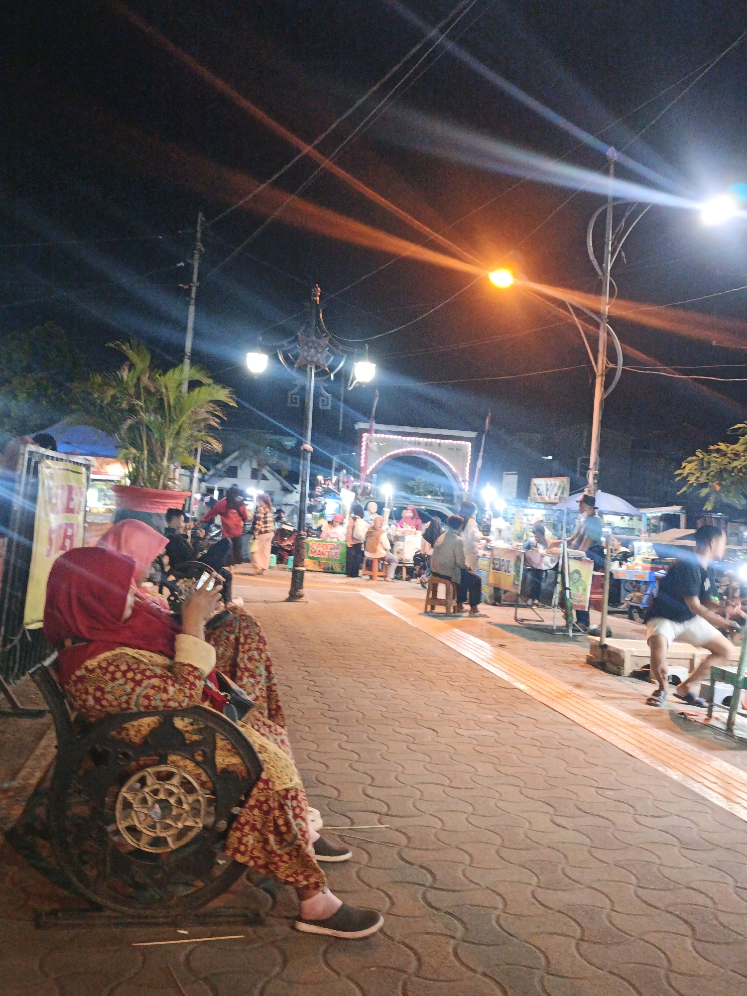 The image depicts a nighttime street scene in Kauman, Indonesia, likely a bustling marketplace or pedestrian area. In the foreground, a person wearing a red hijab and a patterned red and gold outfit sits on an ornate, dark metal bench, looking at their phone. Their lower legs and feet are visible, with the person wearing dark slip-on shoes. The bench has a decorative, spoked wheel-like design on its side.
The background reveals a street lined with various stalls or shops, illuminated by bright streetlights and the warm glow of lamps from the vendors. Several people are visible, some standing, others seated, engaged in shopping or socializing. Arcades or arched structures are present in the distance, adding to the architectural character of the location. The street is paved with textured interlocking tiles. Overhead, electrical wires crisscross the dark sky, illuminated by the artificial lights, creating streaks of light in the photograph. Palm trees and other foliage are visible, indicating a tropical or subtropical environment. The overall atmosphere is lively and indicative of a popular evening gathering spot.