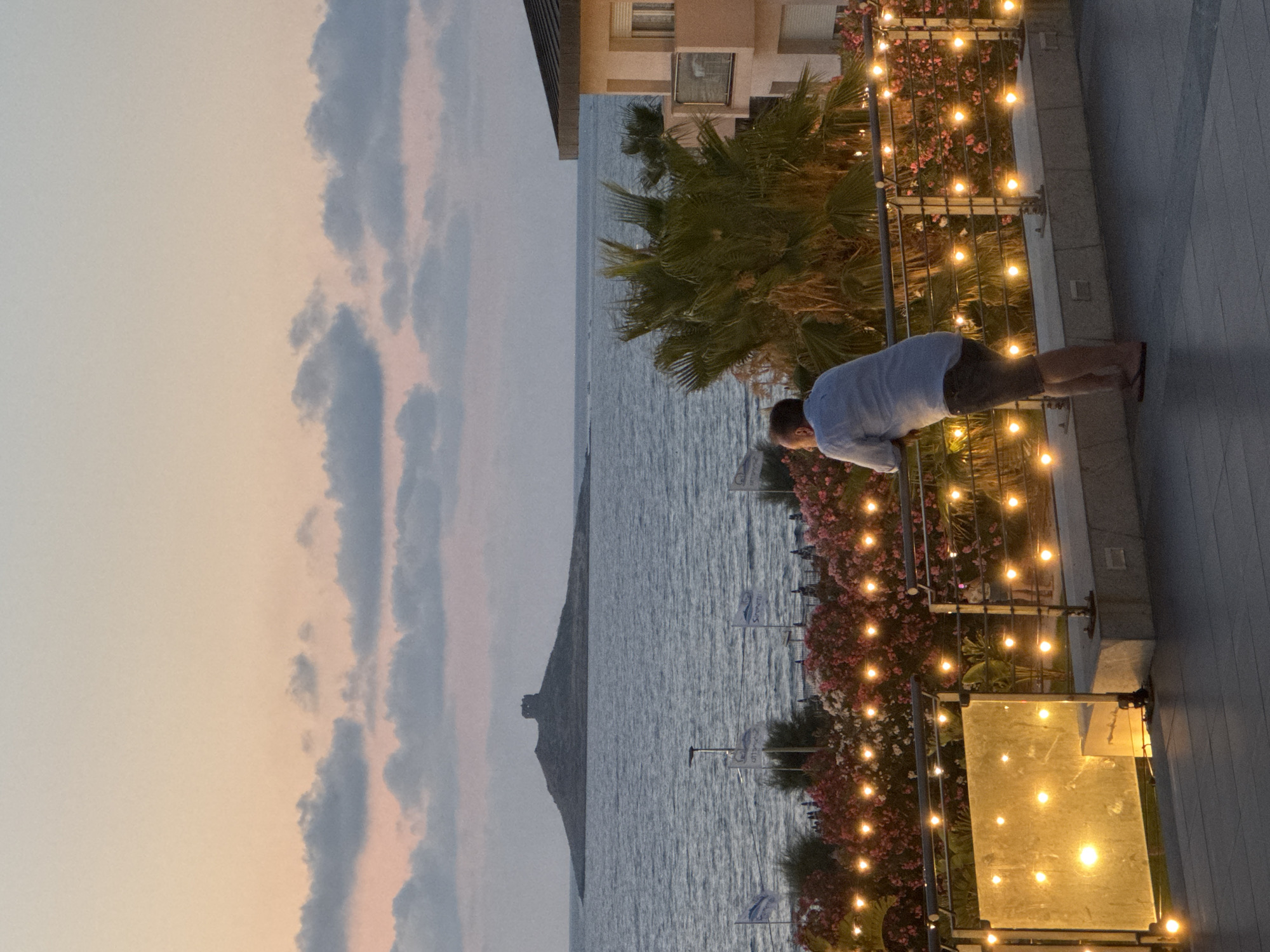 This is a vertical, medium-shot photograph taken at dusk in Isola delle Femmine, Italy. The scene depicts a person leaning over a railing, gazing out at the sea. The sky is a soft gradient of pastel colors, transitioning from pale pink to light blue, with wispy clouds. The sea is calm, reflecting the twilight sky. In the distance, a prominent rocky outcrop or island with a structure on top is visible.
The foreground features a modern building with a balcony area decorated with a string of warm, incandescent lights. Lush green palm trees and vibrant pink and white flowers line the balcony, creating a festive atmosphere. The person in the image is wearing a light-colored, short-sleeved shirt and shorts. They are positioned to the right of the frame, facing away from the camera and towards the sea. Their posture suggests a moment of quiet contemplation or observation.
The lighting indicates it is likely evening or early morning. The overall mood is serene and peaceful, with a hint of the romantic ambiance created by the decorative lights and the natural beauty of the coastal landscape. There are small flags with blue text visible in the distance, but the text is not legible. The image captures a tranquil moment by the Mediterranean Sea.
