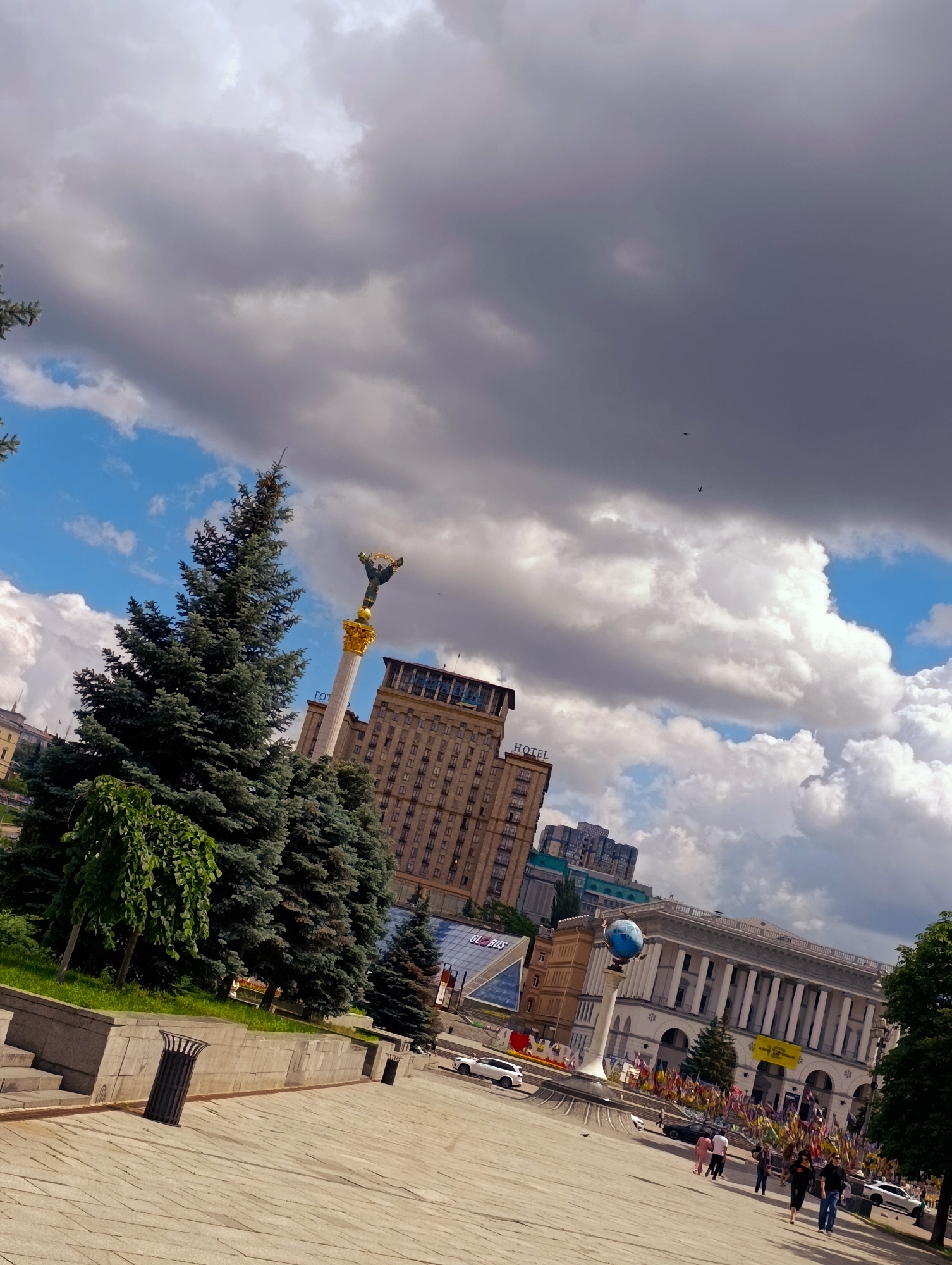 The image depicts Maidan Nezalezhnosti (Independence Square) in Kyiv, Ukraine. In the background, the "Hotel Ukraina" building stands prominently, with its distinctive neoclassical architecture. To its left, the Independence Monument, a tall column topped with a statue of Berehynia, is visible. Several large evergreen trees are in the foreground, obscuring part of the monument and the hotel. The sky is partly cloudy, with large, dramatic grey clouds mixed with patches of bright blue sky. In the middle ground, a grand building with a colonnade is visible, adorned with numerous colorful flags. A spherical blue globe sculpture on a pedestal is also present in front of this building. The foreground is a paved pedestrian area with steps and a low stone wall. Two black trash receptacles are positioned along the wall. Several people are walking in the middle ground and further back, towards the colonnaded building. White cars are parked or driving on a road to the right of the central area. The overall scene suggests a public gathering or event, given the abundance of flags.