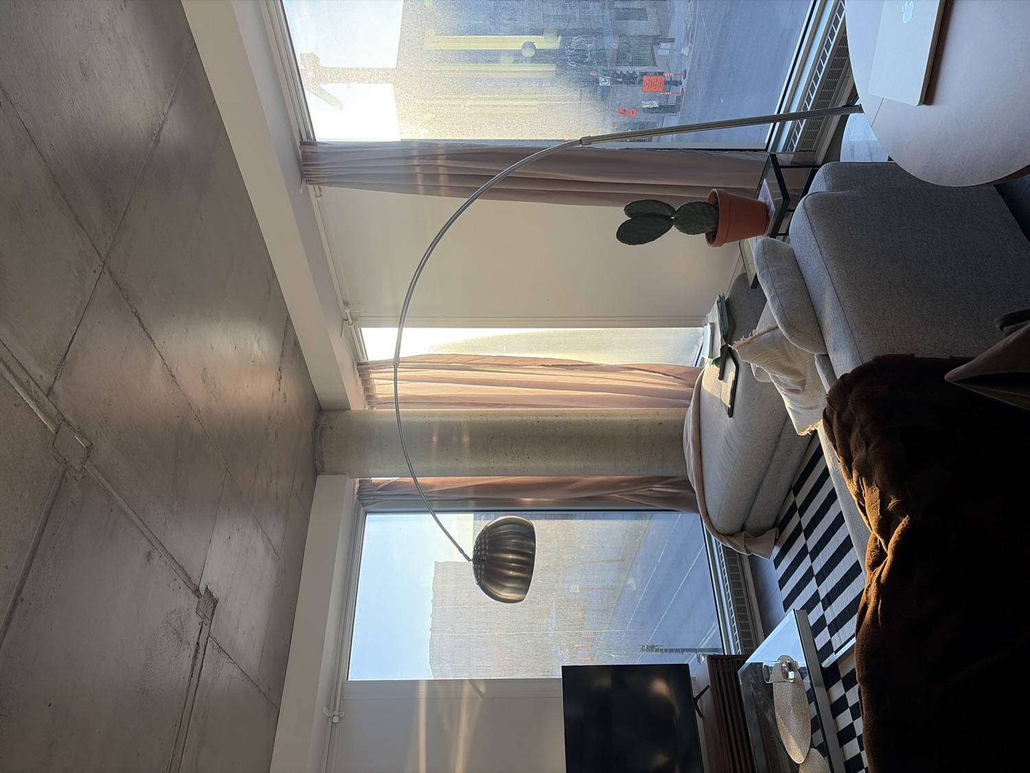 This media file captures a modern, sun-drenched living room interior, likely an apartment in an urban setting such as Montreal. The perspective is looking towards a wall dominated by large, floor-to-ceiling windows that offer a glimpse of city buildings and a street scene outside.
On the left, a raw, textured concrete wall and ceiling with visible panel lines and a prominent concrete pillar establish an industrial-chic aesthetic. Strong sunlight casts distinct shadows across these surfaces.
The right side of the room features a comfortable living area. A light grey sofa, possibly a sectional, is adorned with a brown furry throw blanket. In front of the sofa is a glass coffee table resting on a black and white striped rug. A tall, silver, arched floor lamp with a distinct metallic lampshade extends over this area. Beside the sofa, a small round side table holds a laptop.
Near the windows, between the sofa and the wall, a potted cactus (likely an Opuntia species) sits on a small black table, basking in the sunlight. Light tan curtains are partially drawn along the windows, filtering the intense light. A white heating unit runs along the base of the window wall.
The bright daylight indicates it is either morning or late afternoon, with clear weather outside. No people are visible, suggesting a quiet, unoccupied space. There is no discernible text anywhere in the image.