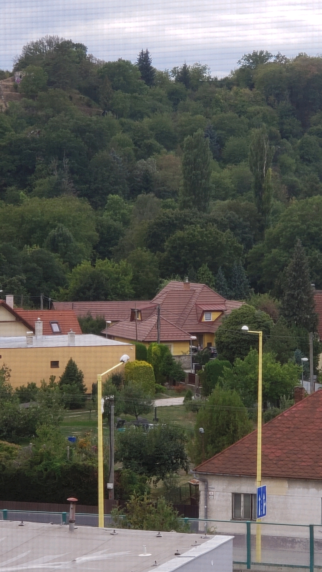 The image shows a residential area with houses and dense greenery, likely a hillside or park in the background. The architecture suggests a Central European setting. Specifically, the image is located in the city of Prešov, Slovakia, and depicts houses with red-tiled roofs. A prominent yellow lamppost stands in the foreground, and a blue traffic sign with two figures is visible on a building. The environment is characterized by a mix of residential structures and abundant trees, indicating a suburban or semi-rural atmosphere within the city.